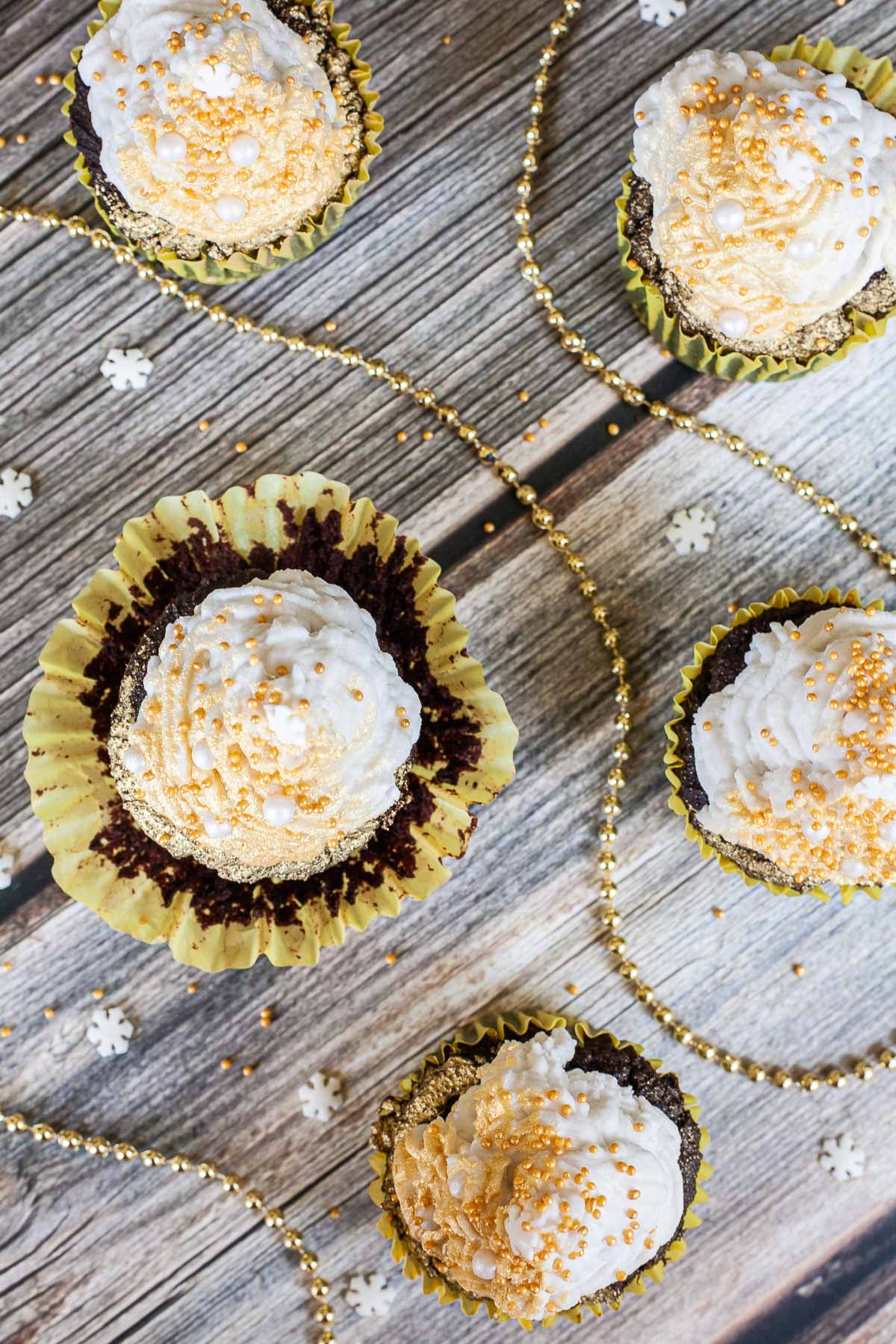 5 dark brownie cupcakes in yellow cupcake liners from above topped with a white frosting sprayed with gold and sprinkled with gold and white pearl sprinkles and a snowflake one on top.