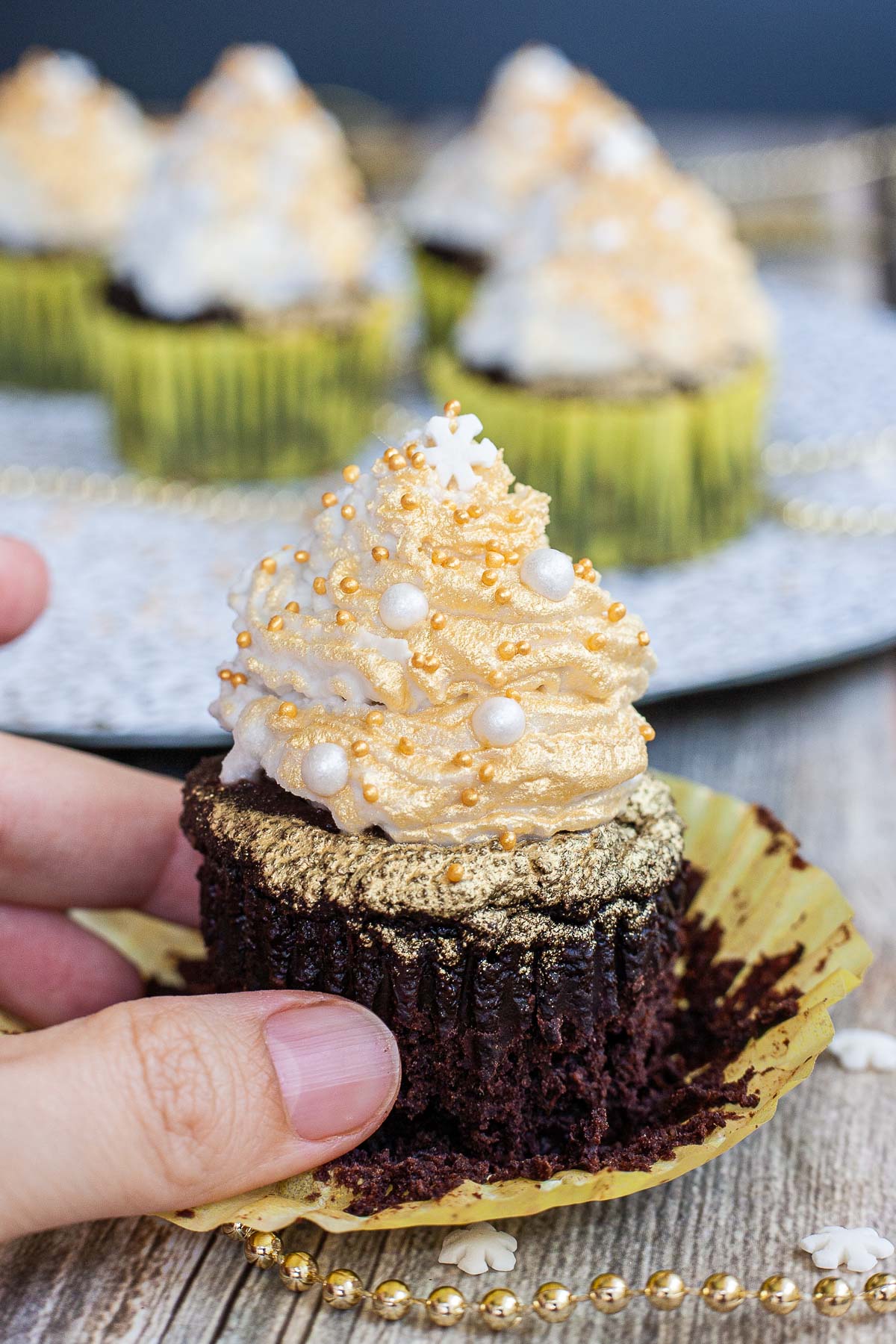 A hand is holding a dark brownie cupcake in yellow unwrapped cupcake liner topped with a white frosting sprayed with gold and sprinkled with gold and white pearl sprinkles and a snowflake one on top. Other cupcakes at the back.