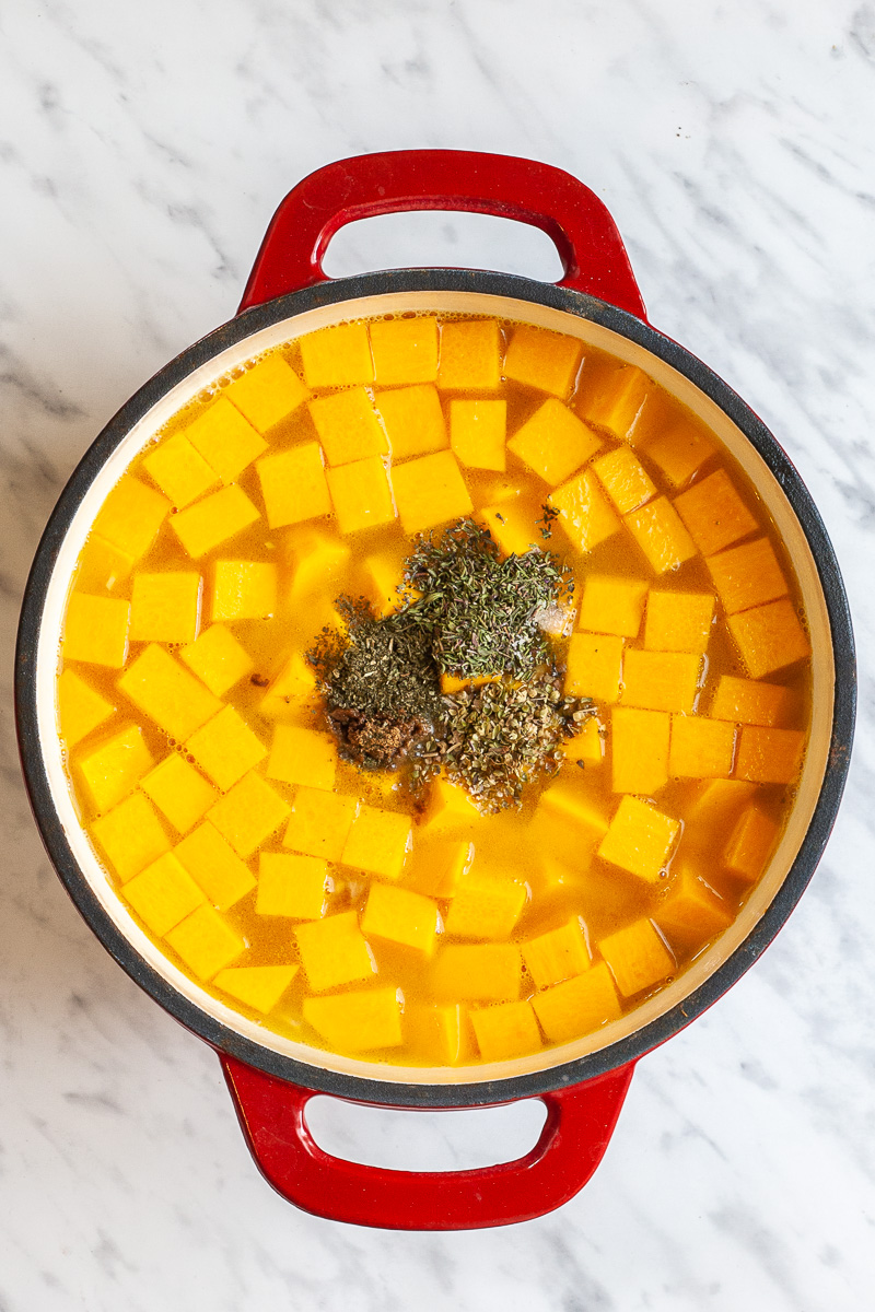 Red white cast iron skillet with broth, orange squash cubes and small heaps of green dried herbs.
