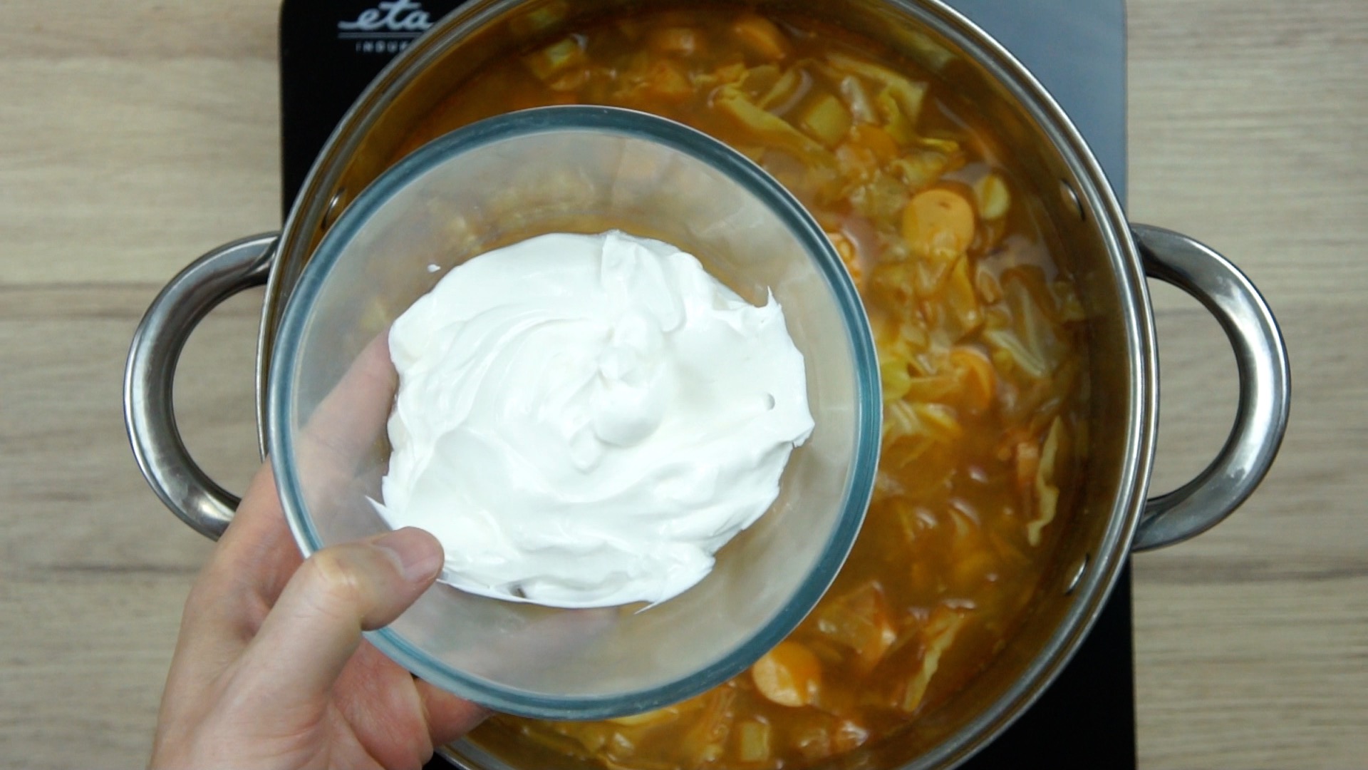 A hand is holding a small glass bowl with white cream above a brown soup with cabbage leaves and other vegetables in a stockpot.