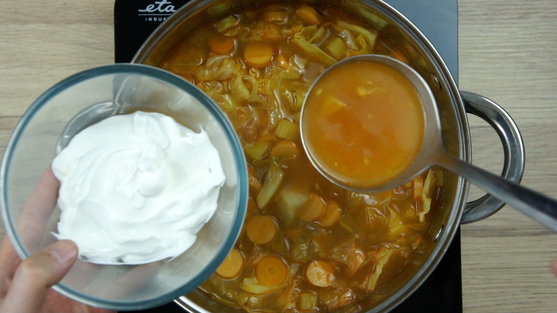 A hand is holding a small glass bowl with white cream above a brown soup with cabbage leaves and other vegetables in a stockpot. A ladle is taking some from the soup.