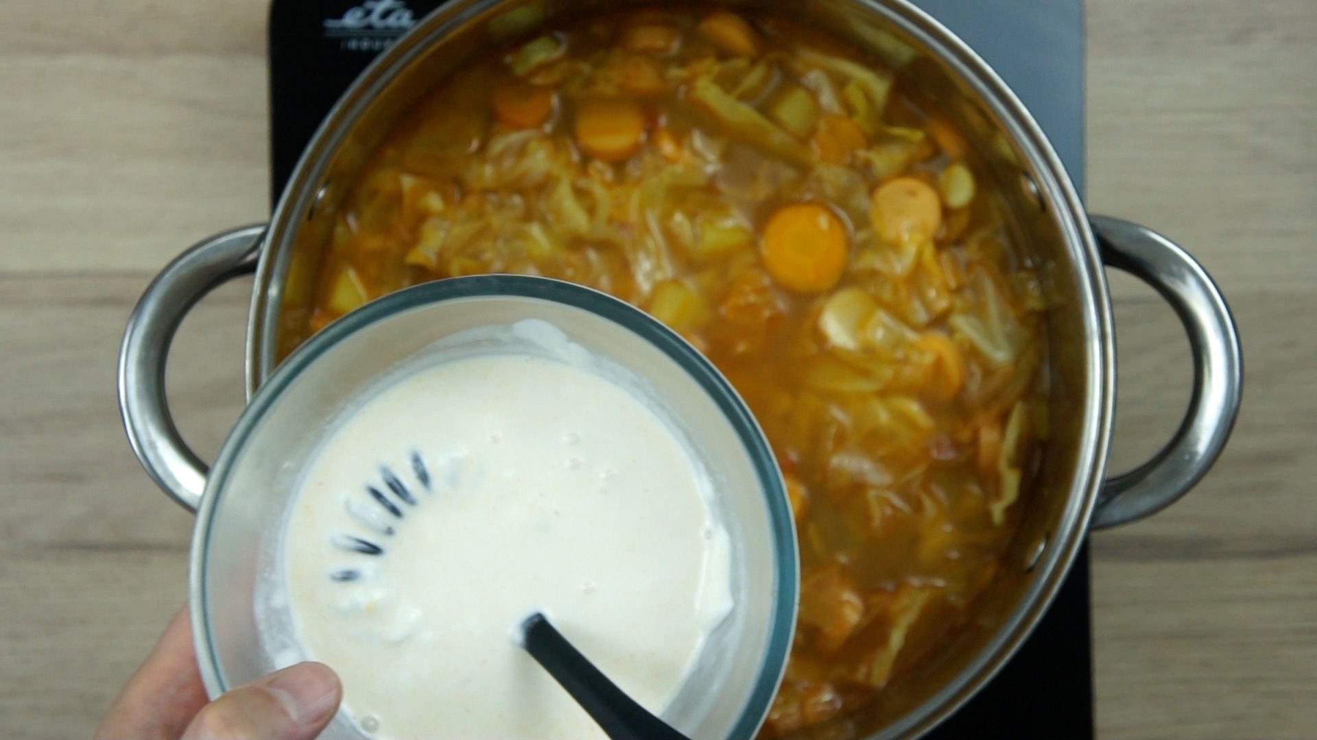 A hand is mixing white cream and some liquid from the soup in a small glass bowl above a brown soup with cabbage leaves and other vegetables in a stockpot.