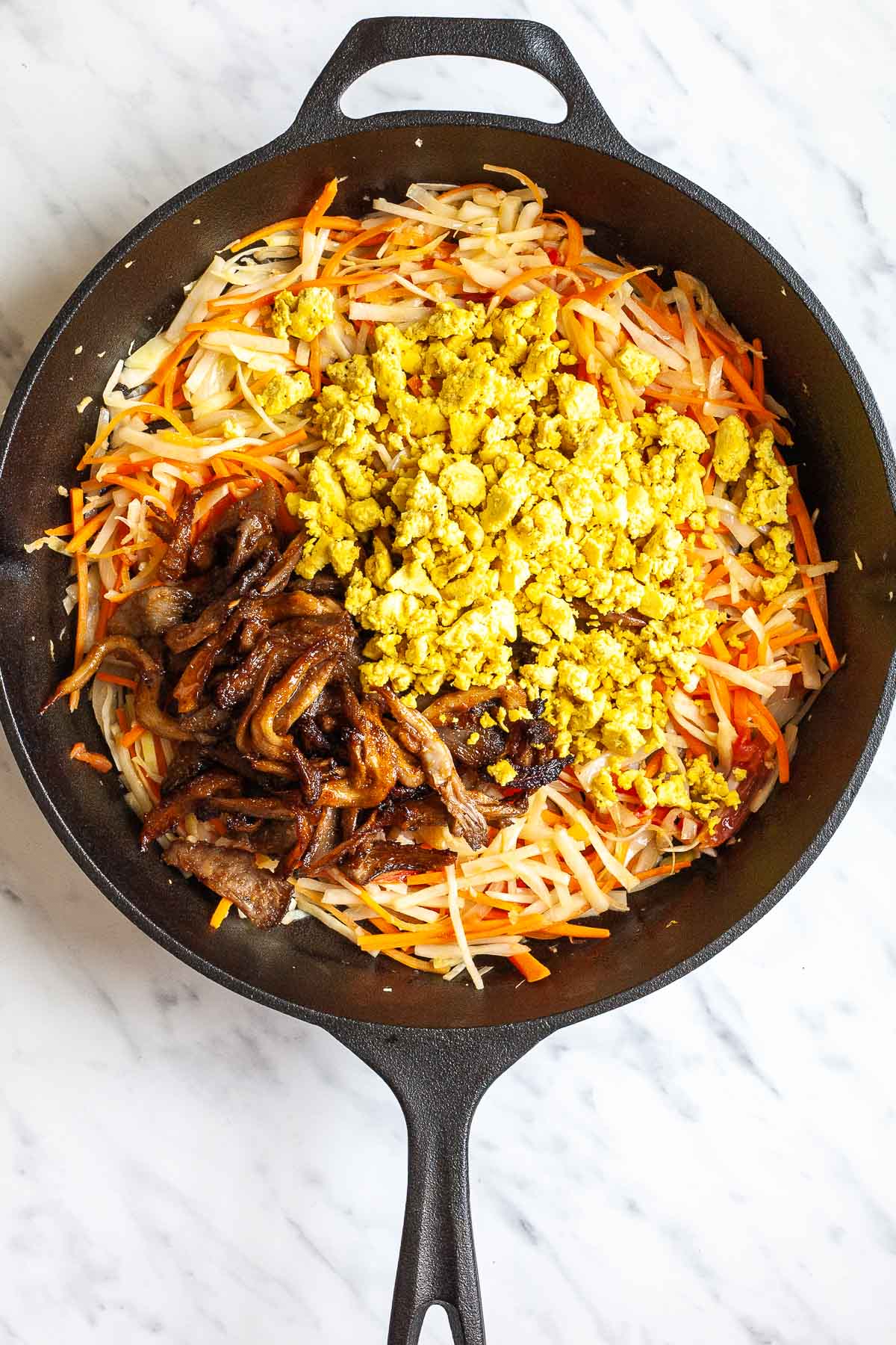 Cast iron skillet with sliced onion, cabbage, tomatoes, and carrots topped with a heap of crispy oyster mushroom shreds and scrambled yellow tofu. 