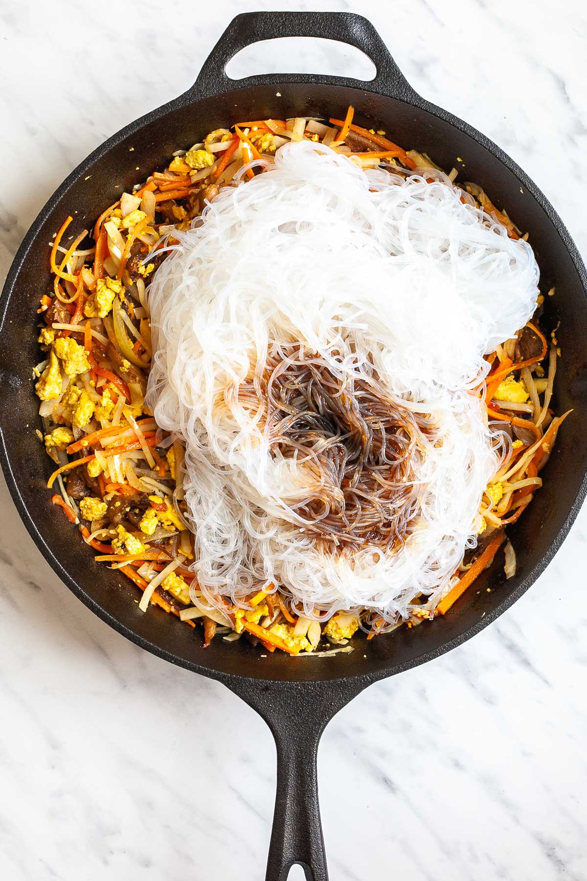 Cast iron skillet with a mix of sliced onion, cabbage, tomatoes, carrots, crispy oyster mushroom shreds and scrambled yellow tofu topped with a heap of thin glass noodles in a bit of dark brown sauce. 
