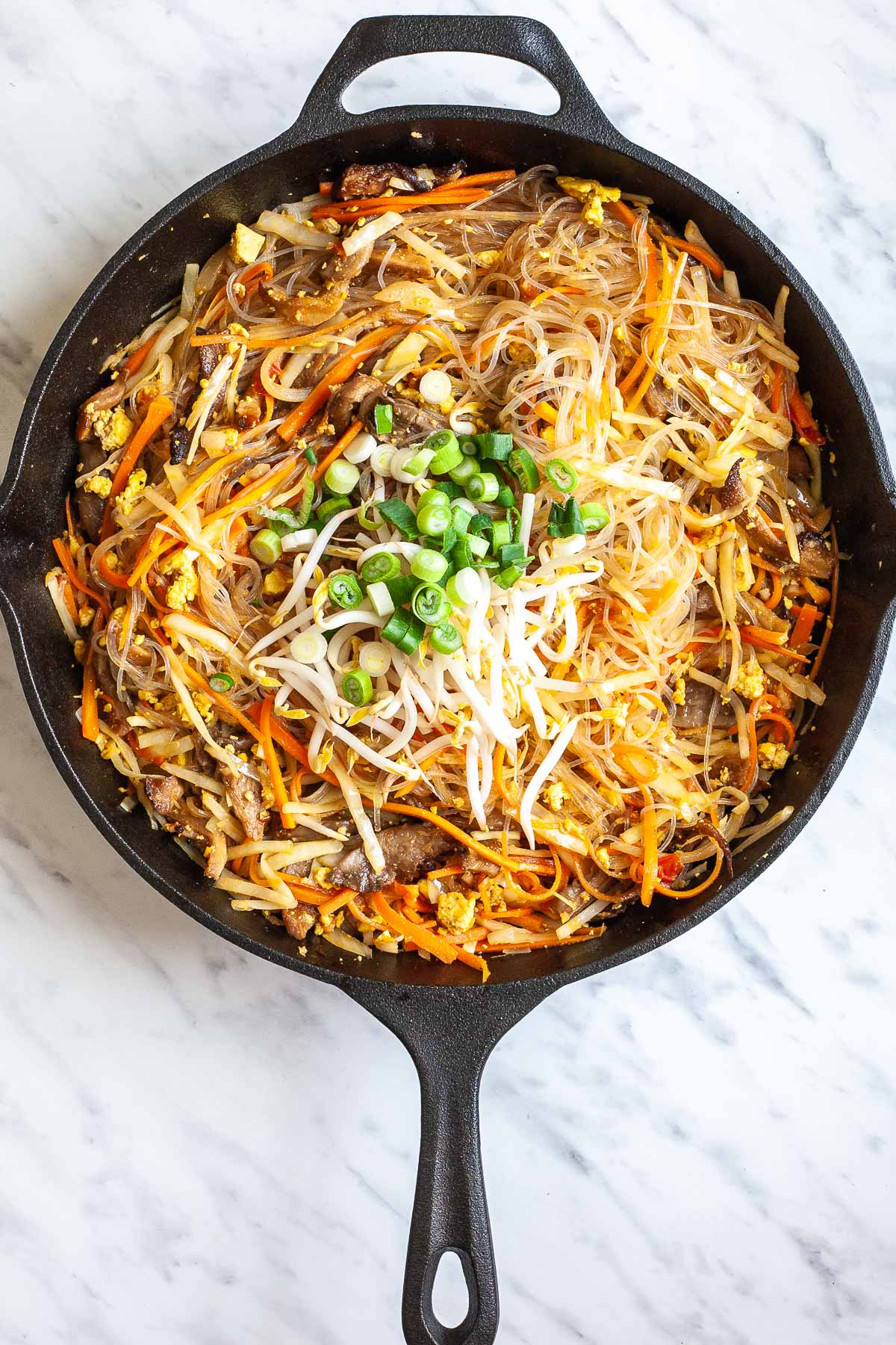 Cast iron skillet with a mix of sliced onion, cabbage, tomatoes, carrots, crispy oyster mushroom shreds, scrambled yellow tofu, and glass noodles  topped with bean sprouts and chopped scallion. 