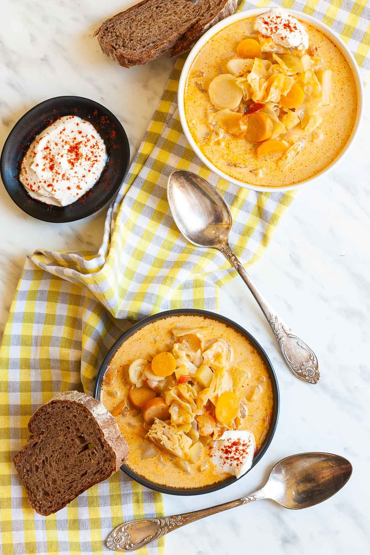 Two bowls with orange soup, sausage, carrot slices, diced potatoes and chopped cabbage topped with white sour cream. A slice of bread is placed next to the bowl as well as two spoons.