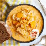 Black bowl with orange soup, sausage, carrot slices, diced potatoes and chopped cabbage topped with white sour cream. A slice of bread is placed next to the bowl as well as two spoons.