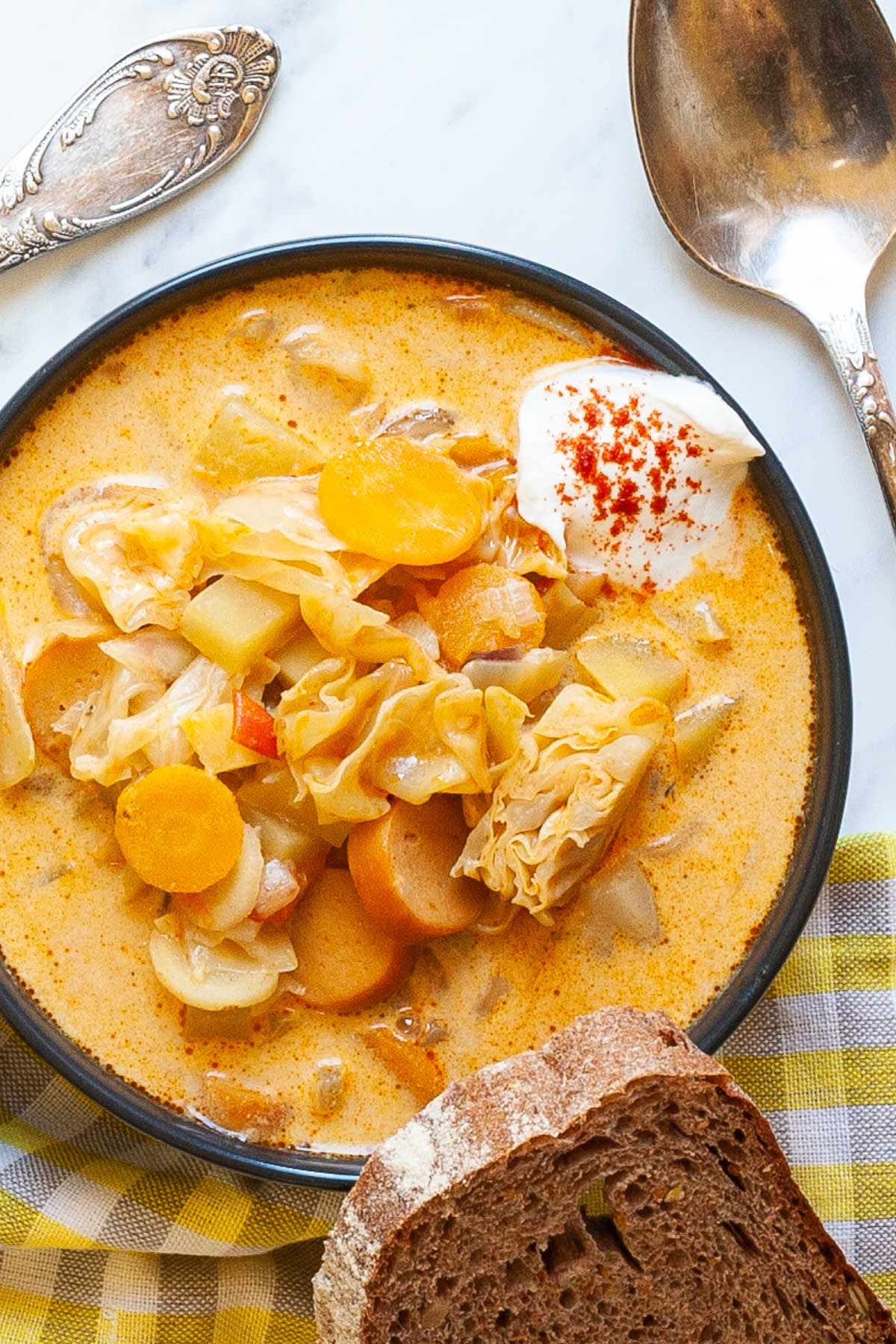 Black bowl with orange soup, sausage, carrot slices, diced potatoes and chopped cabbage topped with white sour cream. A slice of bread is placed next to the bowl as well as two spoons. 