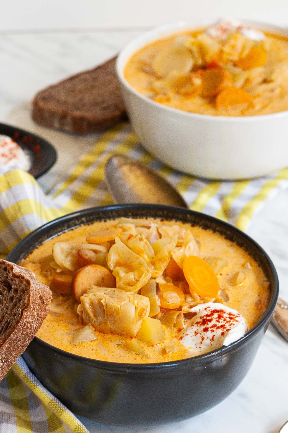 Two bowls with orange soup, sausage, carrot slices, diced potatoes and chopped cabbage topped with white sour cream. A slice of bread is placed next to the bowl as well as two spoons.