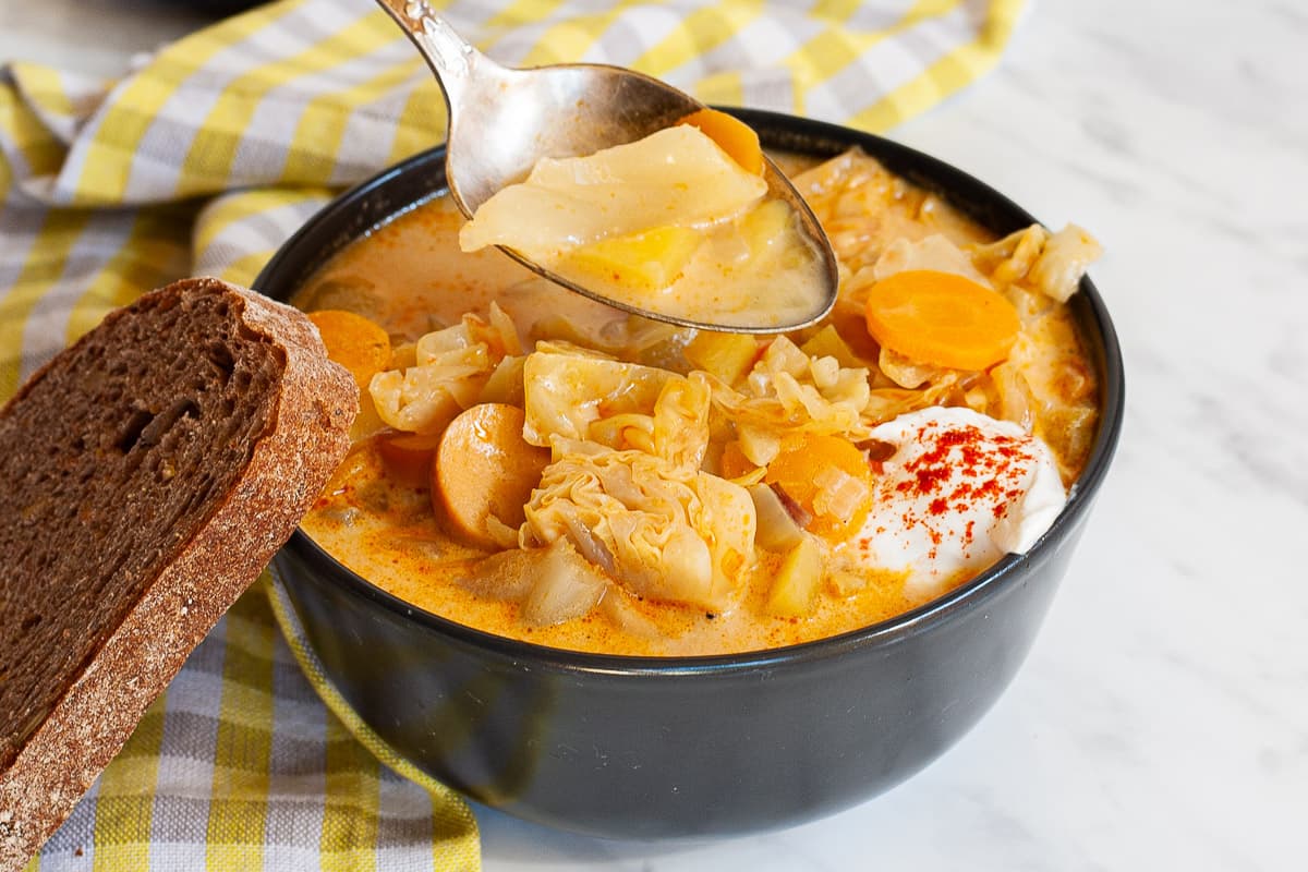 A black bowl of orange soup, sausage, carrot slices, diced potatoes and chopped cabbage topped with white sour cream. A slice of bread is placed next to the bowl. A hand is holding a spoon with some soup.
