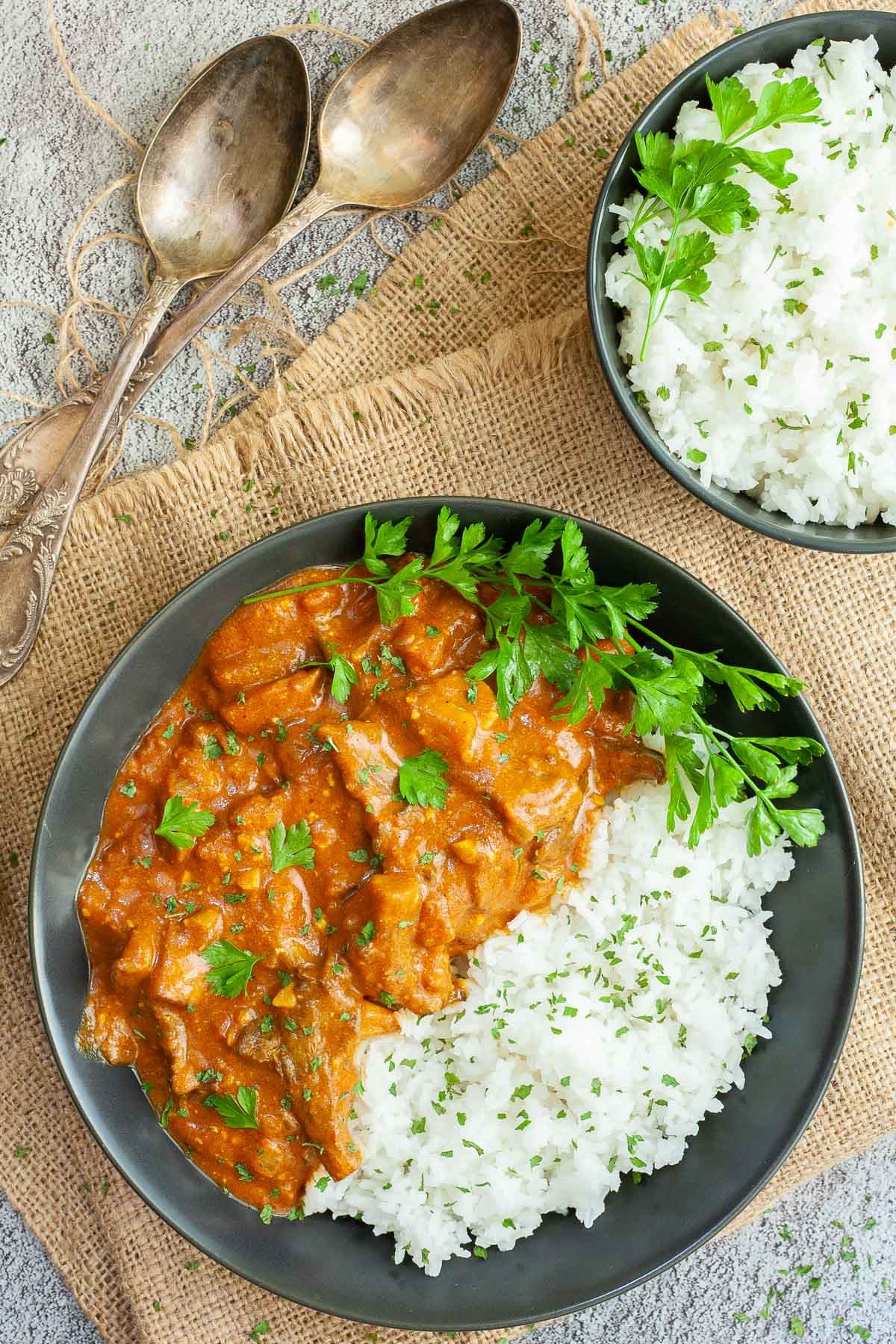 Large black plate with white rice and mushroom shreds in orange thick sauce sprinkled with chopped green herbs and a twig of parsley on top. Two spoons and a small bowl of leftover rice are placed next to it.