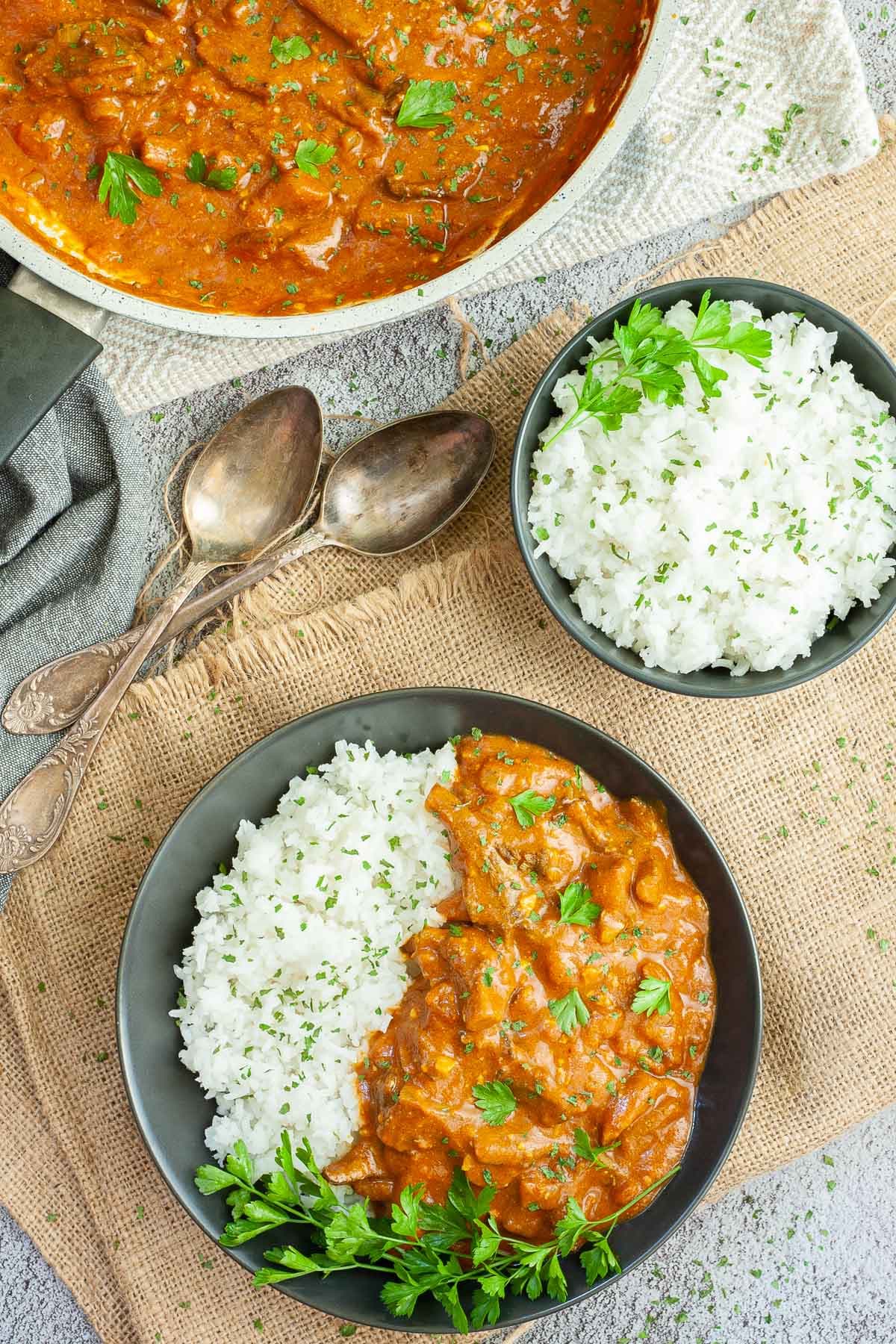 Large black plate with white rice and mushroom shreds in orange thick sauce sprinkled with chopped green herbs and a twig of parsley on top. Two spoons, a frying pan with sauce and a small bowl of leftover rice are placed next to it.