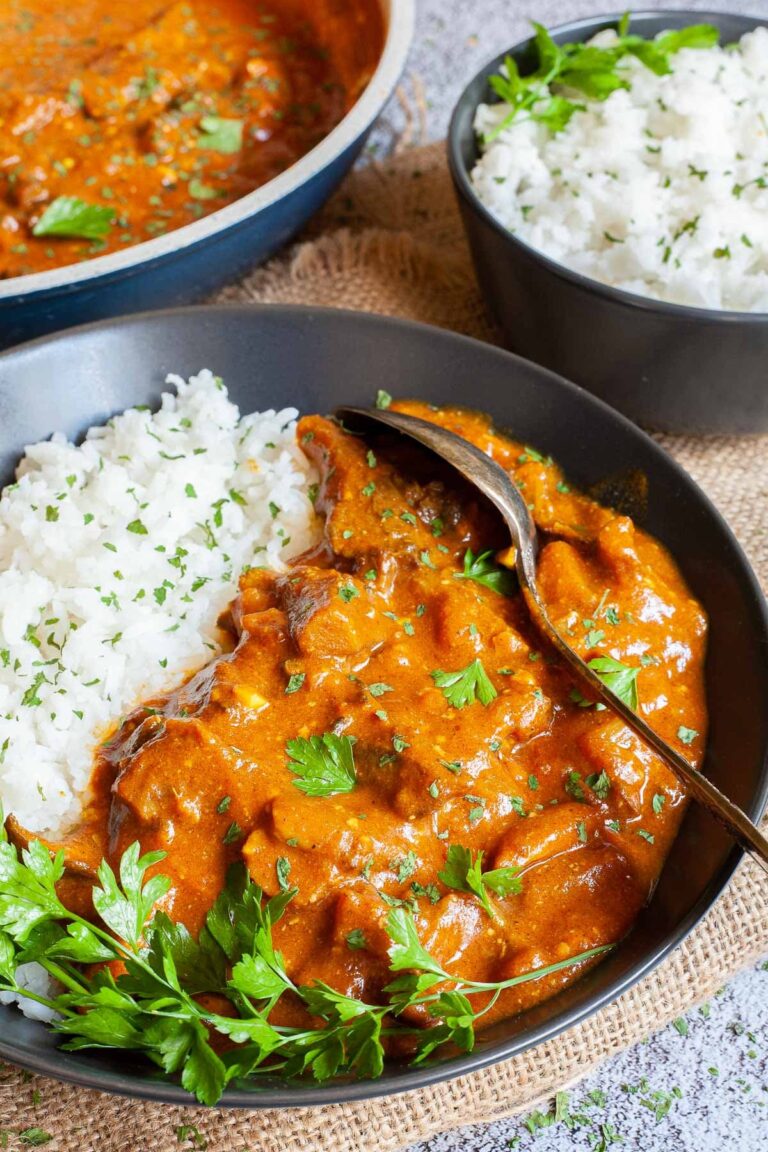 Large black plate with white rice and mushroom shreds in orange thick sauce sprinkled with chopped green herbs and a twig of parsley on top. Two spoons, a frying pan with sauce and a small bowl of leftover rice are placed next to it.