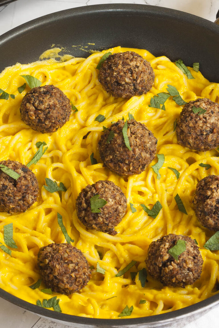 Several mushroom meatballs on top of spaghetti with yellow creamy sauce served on a black plate. A fork is taking on meatballs from the middle.