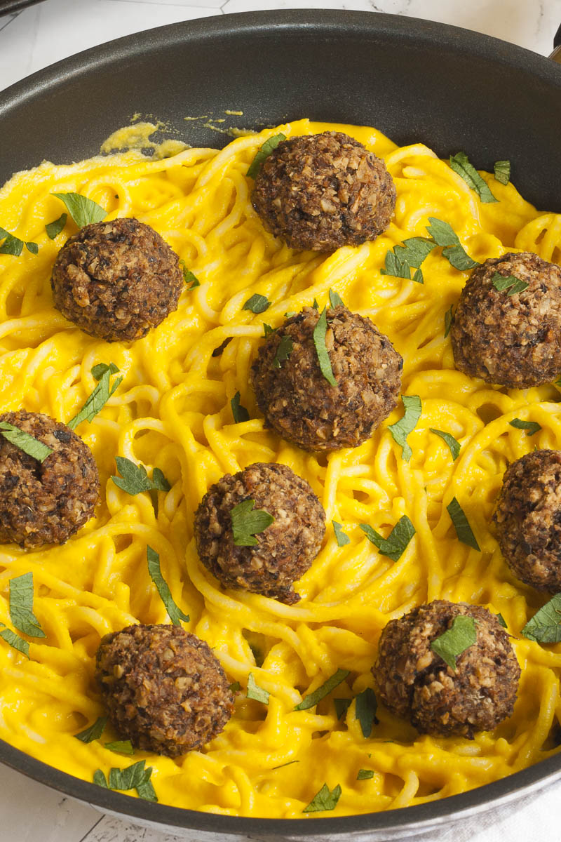 Several mushroom meatballs on top of spaghetti with yellow creamy sauce served on a black plate. A fork is taking on meatballs from the middle.