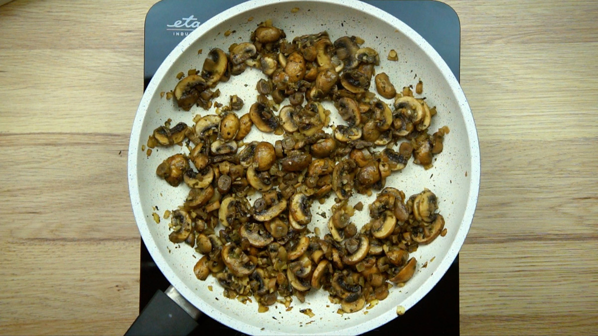 White frying pan with dark brown cooked mushroom slices and chopped onion.