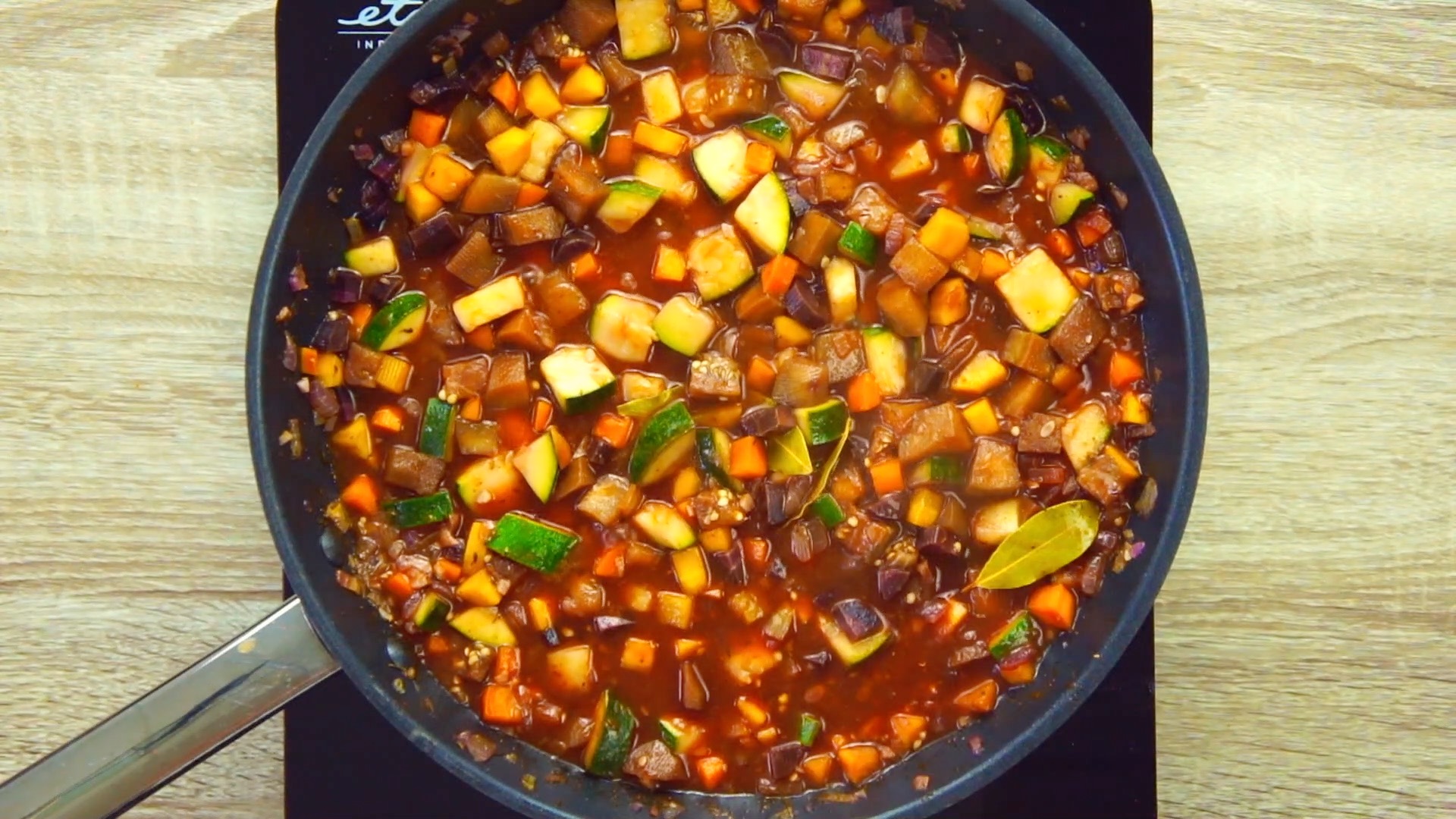 Black frying pan with lots of colorful diced vegetables like eggplant, zucchini, carrots in tomato sauce