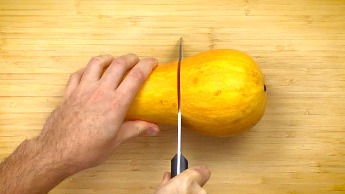 A hand is cutting a peeled butternut squash in the middle. 