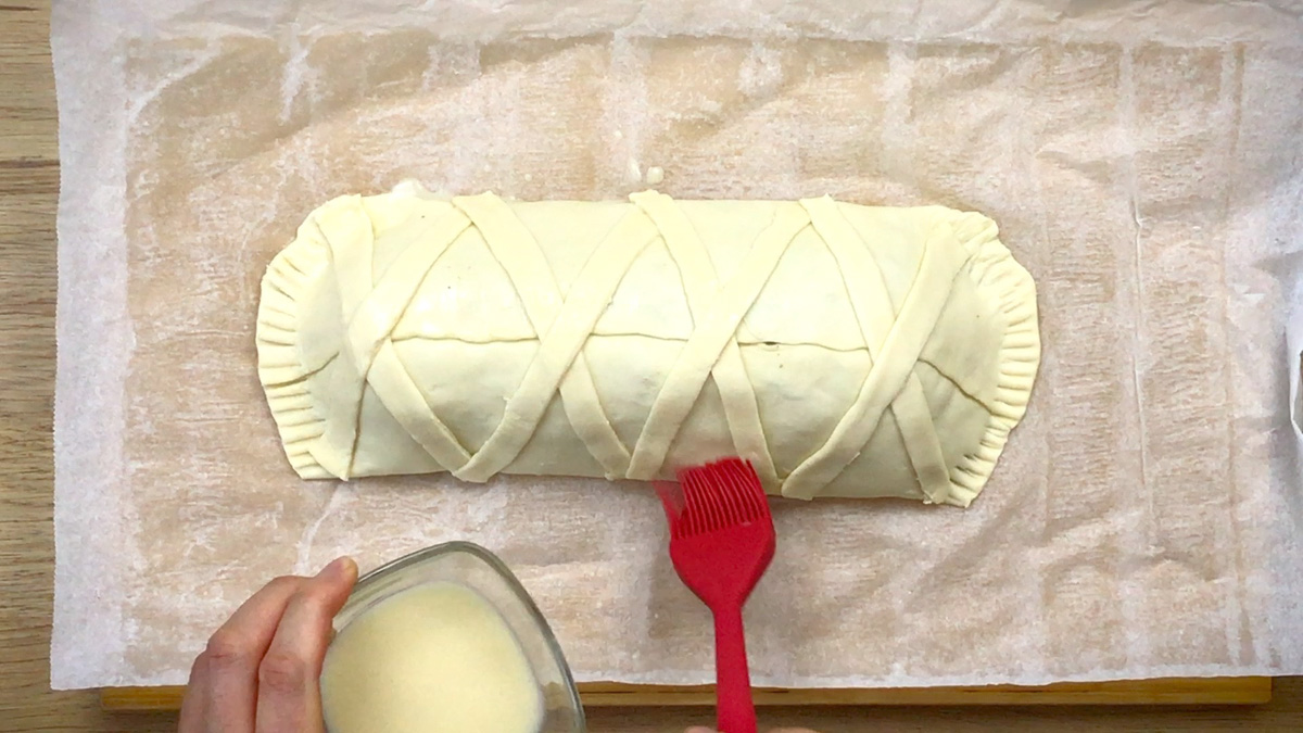 A parchment paper with a rectangle-shaped filled puff pastry. It is decorated by crossing puff pastry strips. A hand is brushing it with milk using a red brush. 