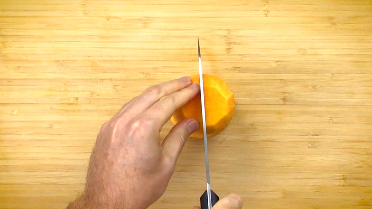 A hand is cutting the neck of a peeled butternut squash into two. 