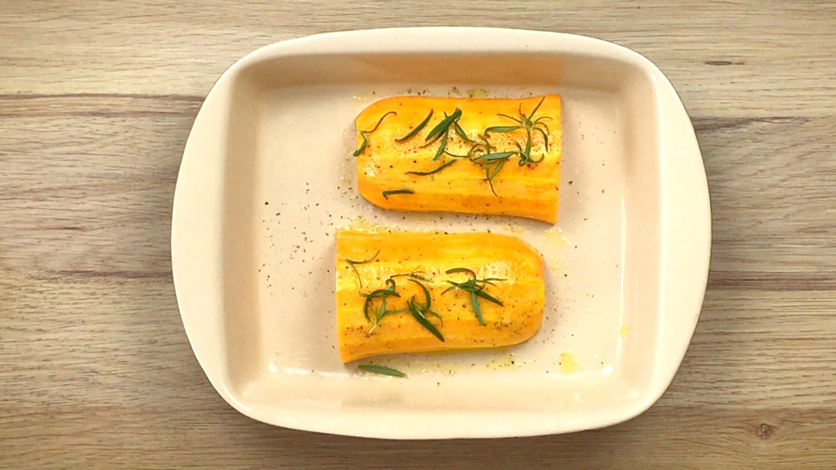 Two halves of the butternut squash neck is placed in an oven-safe ceramic dish and sprinkled with fresh rosemary leaves. 