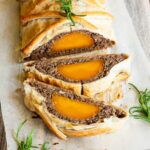 2 slices of vegan wellington on the side. Yellow squash with brown puree around wrapped in a pastry shell. Green herbs are around.