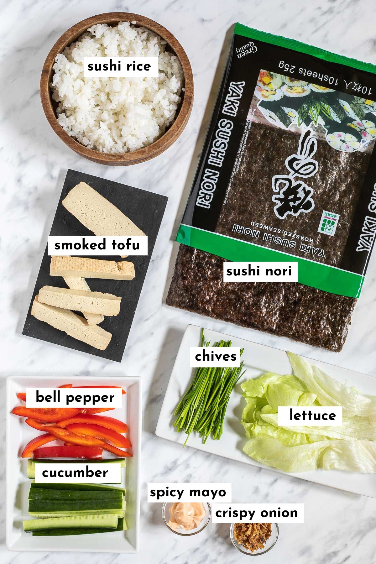 Ingredients of tofu sushi like rice in a wooden bowl, smoked tofu slices on a black plate, cucumber and red pepper strips, lettuce, chives, nori sheets, crispy fried onion pieces, red orange mayo in a small glass bowl