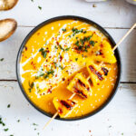Yellow cream soup in a black bowl topped with charred pineapple wedges, drizzled with a white sauce and sprinkled with chopped green herbs, red flakes and sesame seeds.