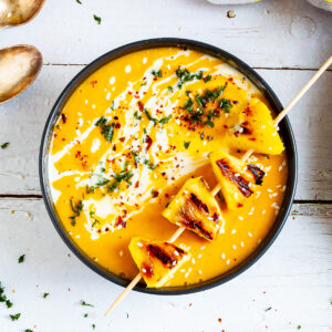 Yellow cream soup in a black bowl topped with charred pineapple wedges, drizzled with a white sauce and sprinkled with chopped green herbs, red flakes and sesame seeds.