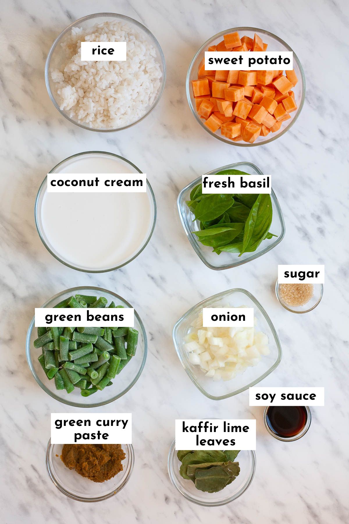 Ingredients of Thai green curry fried rice placed in small glass bowls like rice, sweet potato cubes, green bean pieces, coconut cream, sugar, kaffir lime leaves, basil leaves, onion, soy sauce, and green curry paste