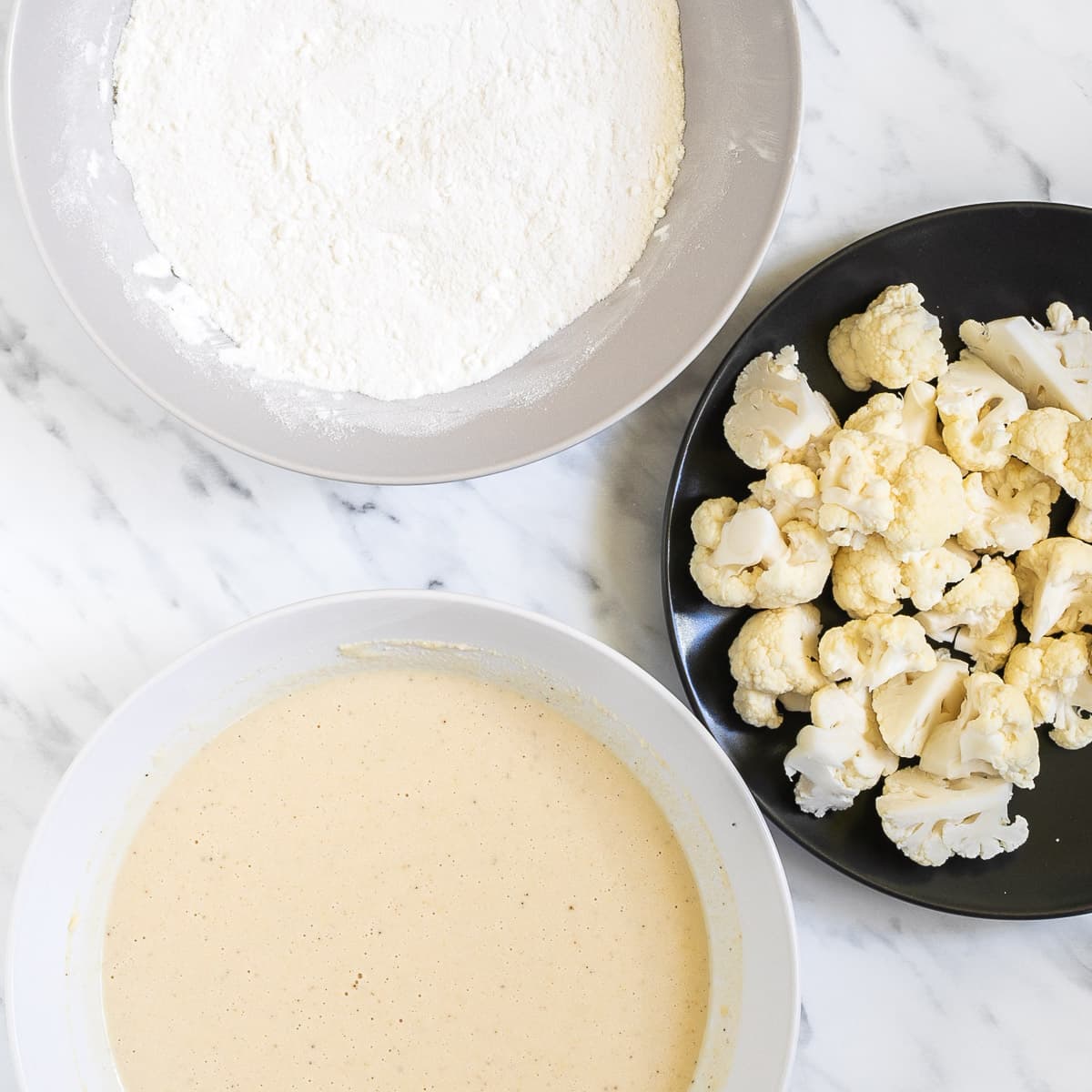 One white bowl of white flour, one white bowl of light yellow batter and one black bowl of breaded cauliflower florets.