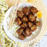 Sticky brown breaded cauliflower florets are served on top of rice in a white bowl. It is sprinkled with chopped chives and sesame seeds. Chopsticks are placed on top of the rice.
