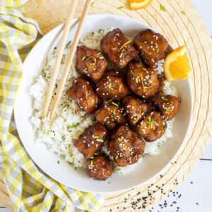 Sticky brown breaded cauliflower florets are served on top of rice in a white bowl. It is sprinkled with chopped chives and sesame seeds. Chopsticks are placed on top of the rice.