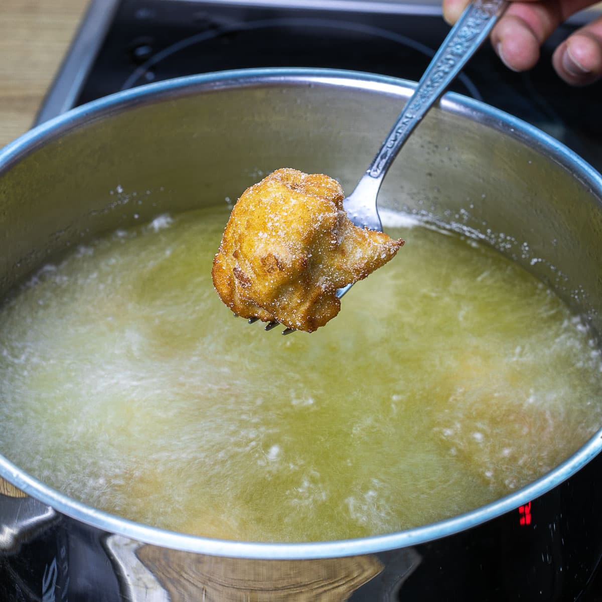 A hand is holding a fork with a crispy breaded deep-fried cauliflower floret right above a deep pan with hot oil.
