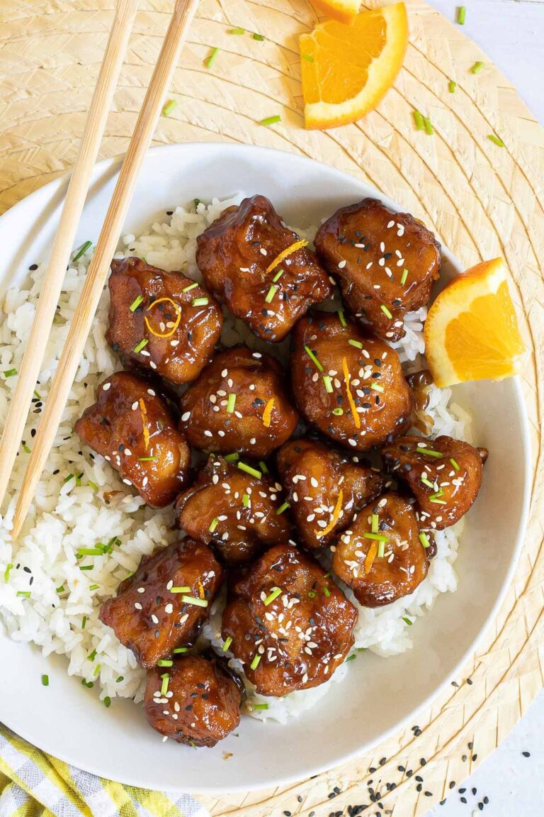 Sticky brown breaded cauliflower florets are served on top of rice in a white bowl. It is sprinkled with chopped chives and sesame seeds. Chopsticks are placed on top of the rice.