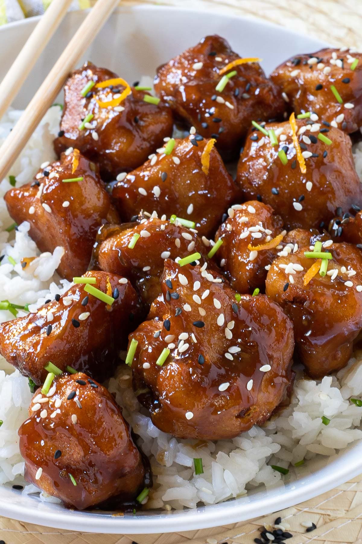 Sticky brown breaded cauliflower florets are served on top of rice in a white bowl. It is sprinkled with chopped chives and sesame seeds. Chopsticks are placed on top of the rice.