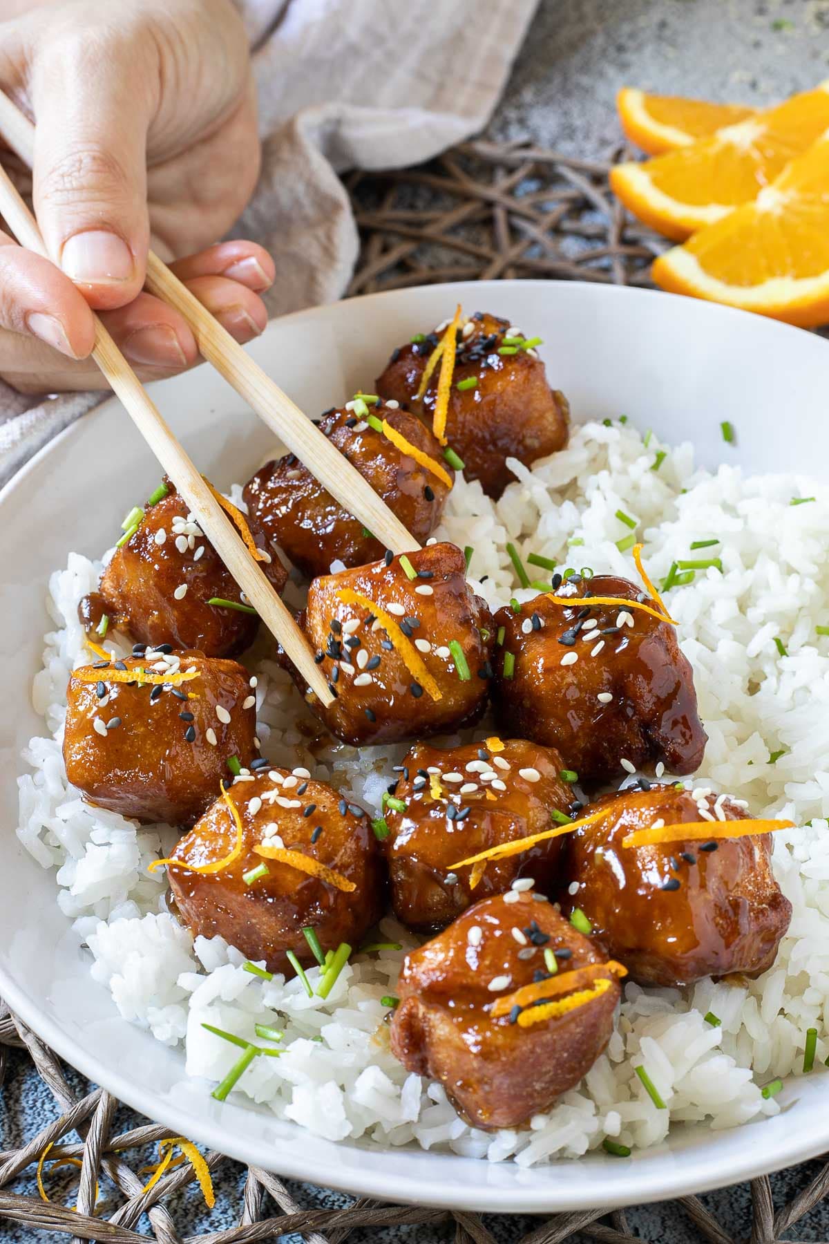 Sticky brown breaded tofu cubes are served on top of rice in a white bowl. It is sprinkled with chopped chives and sesame seeds. A hand is holding chopsticks to take one.