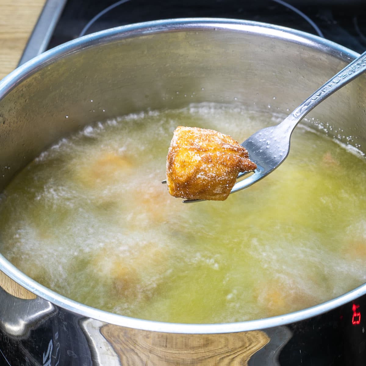 A hand is holding a fork with a crispy breaded deep-fried tofu cube right above a deep pan with hot oil.