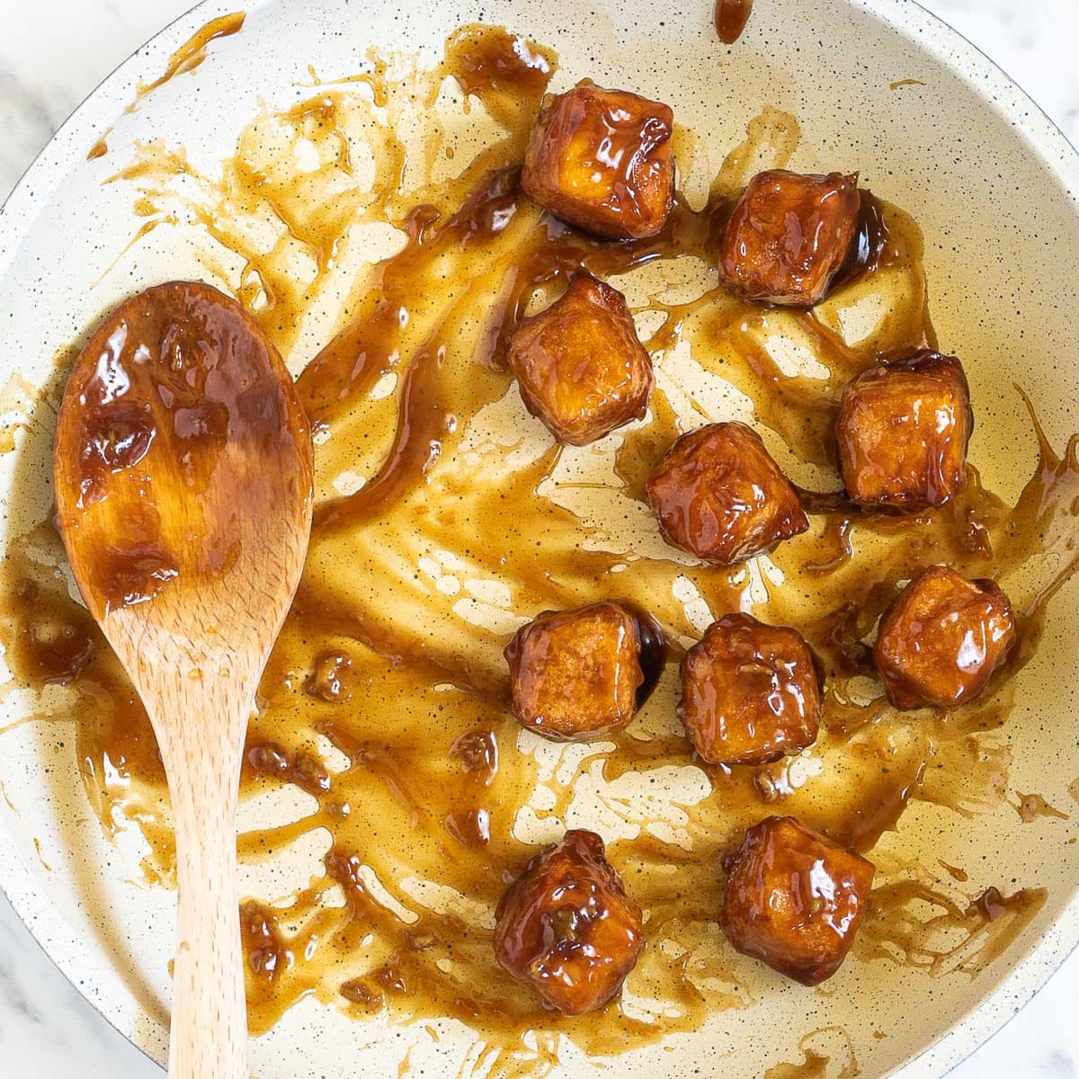 A white frying pan with a dark brown sticky sauce and several breaded crispy tofu cubes. A wooden spatula which was used to glaze the cubes are placed next to them.