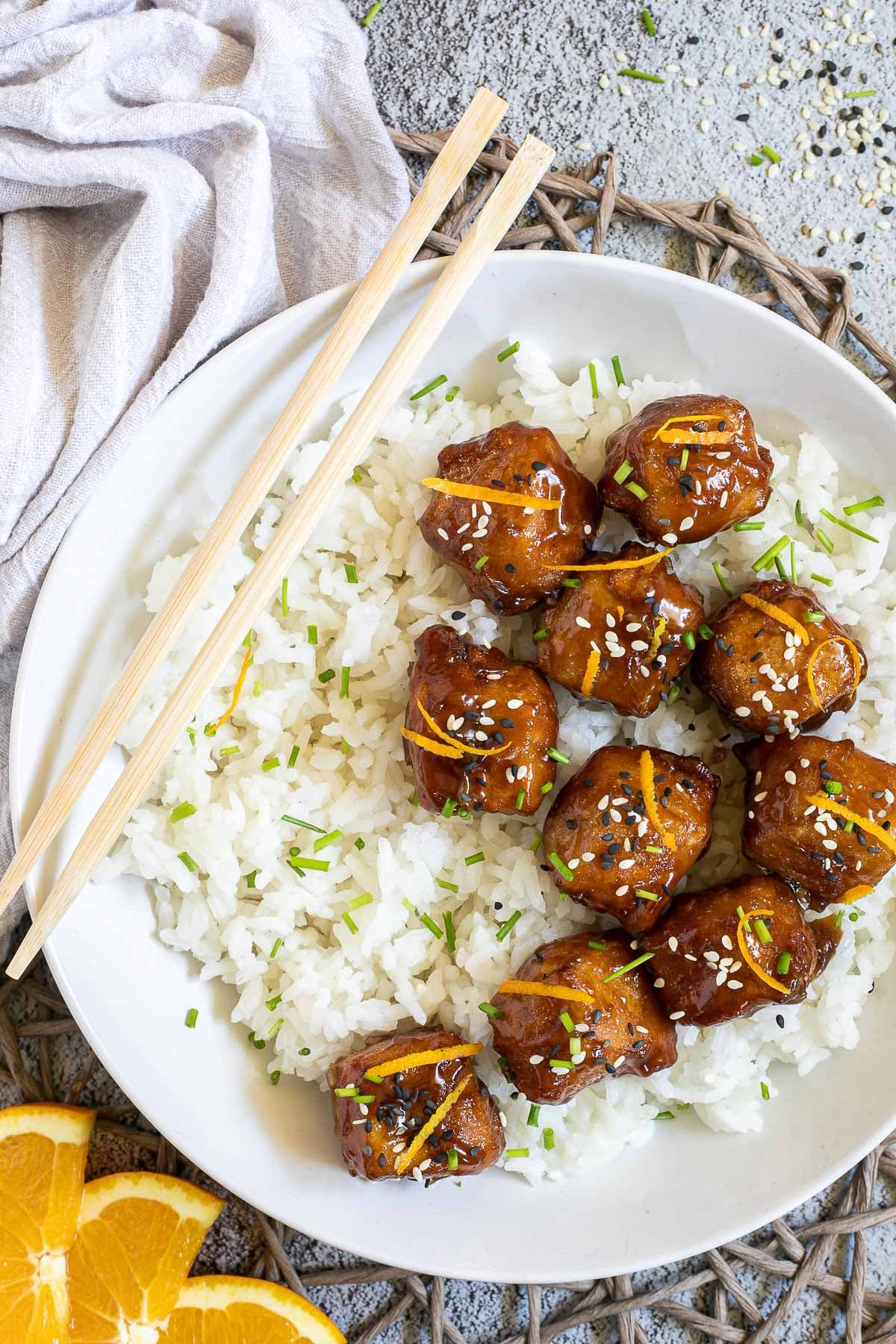 Sticky brown breaded tofu cubes are served on top of rice in a white bowl. It is sprinkled with chopped chives and sesame seeds. Chopsticks are placed on the rice.