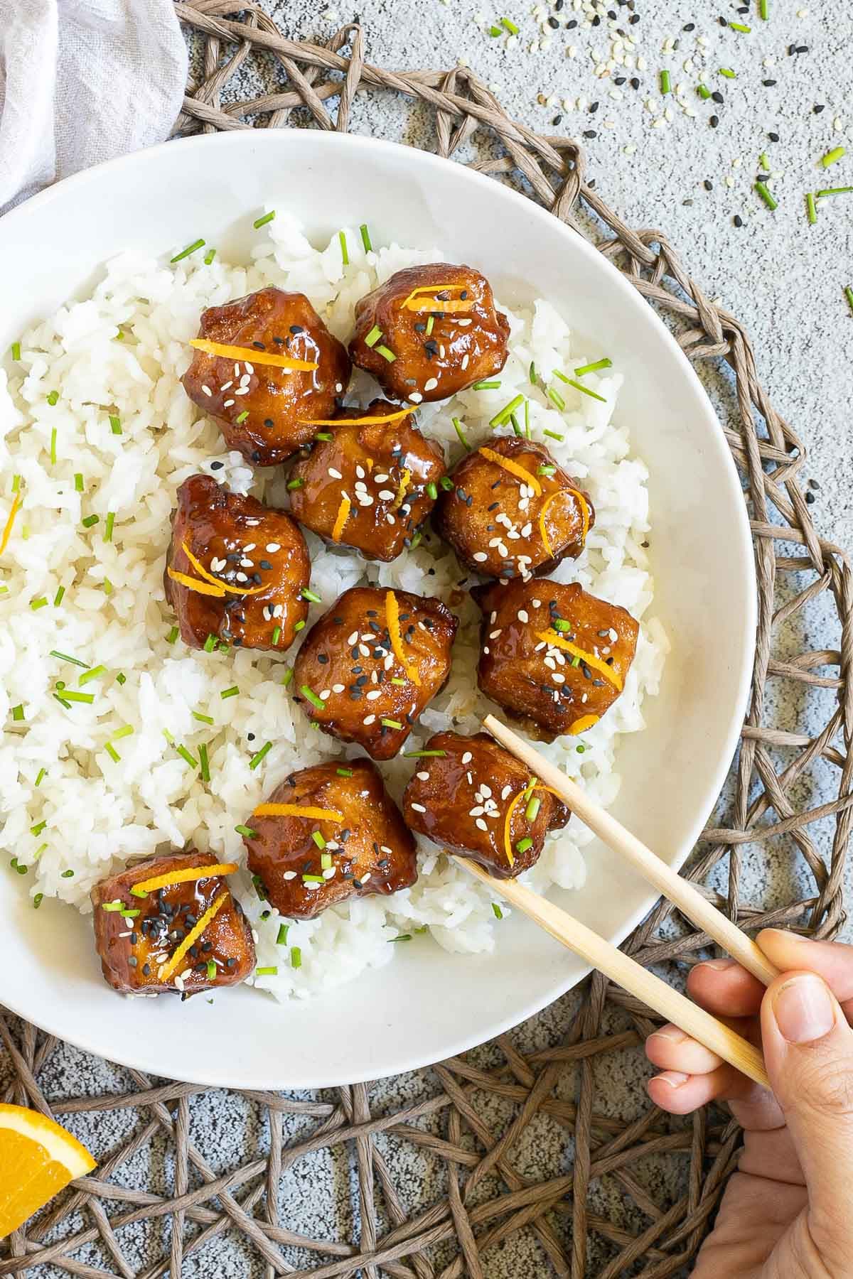 Sticky brown breaded tofu cubes are served on top of rice in a white bowl. It is sprinkled with chopped chives and sesame seeds. A hand is holding chopsticks to take one.