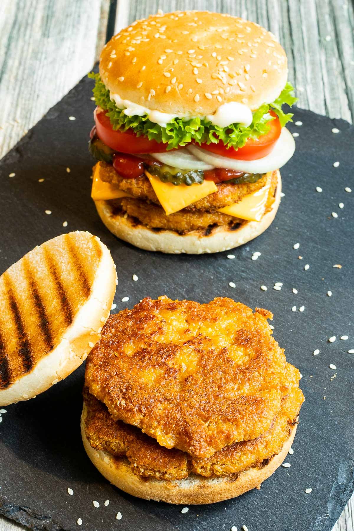 2 burgers on a black board. One with two patties, cheese in between, pickled cucumber slices, onion slices, tomato and ruffled lettuce. And the other is with 2 patties and nothing else, not even top bun.