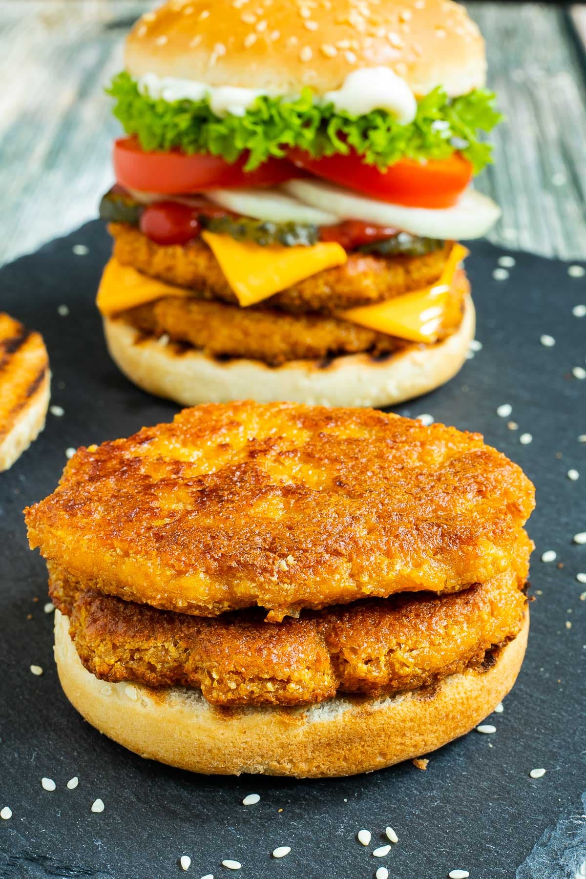 2 burgers on a black board. One with two patties, cheese in between, pickled cucumber slices, onion slices, tomato and ruffled lettuce. And the other is with 2 patties and nothing else, not even top bun.