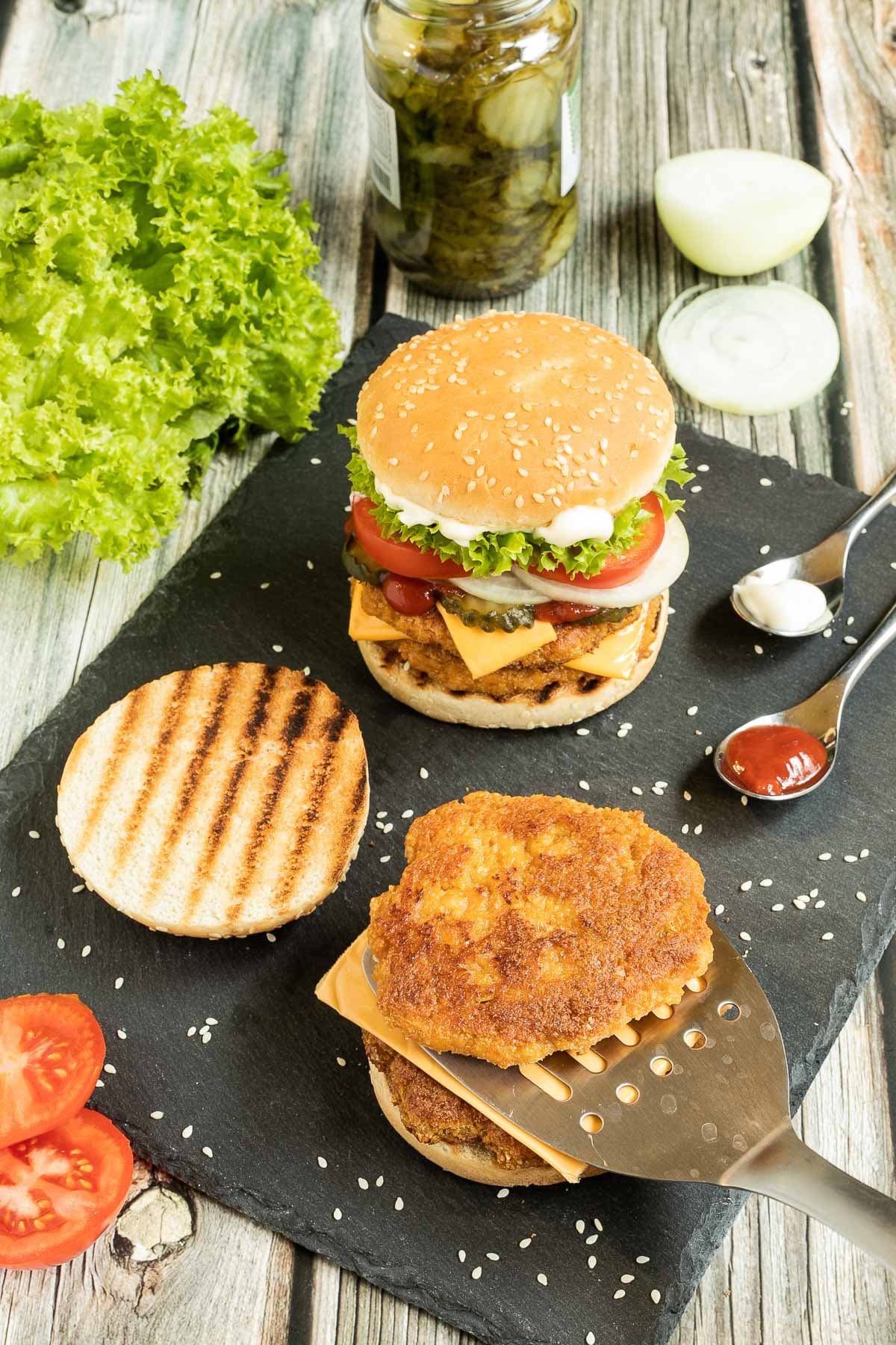 2 burgers on a black board. One with two patties, cheese in between, pickled cucumber slices, onion slices, tomato and ruffled lettuce. And the other is with 2 patties and nothing else, not even top bun. Leftover ingredients are around it like lettuce, jar of pickled cucumber, onion, and tomatoes.
