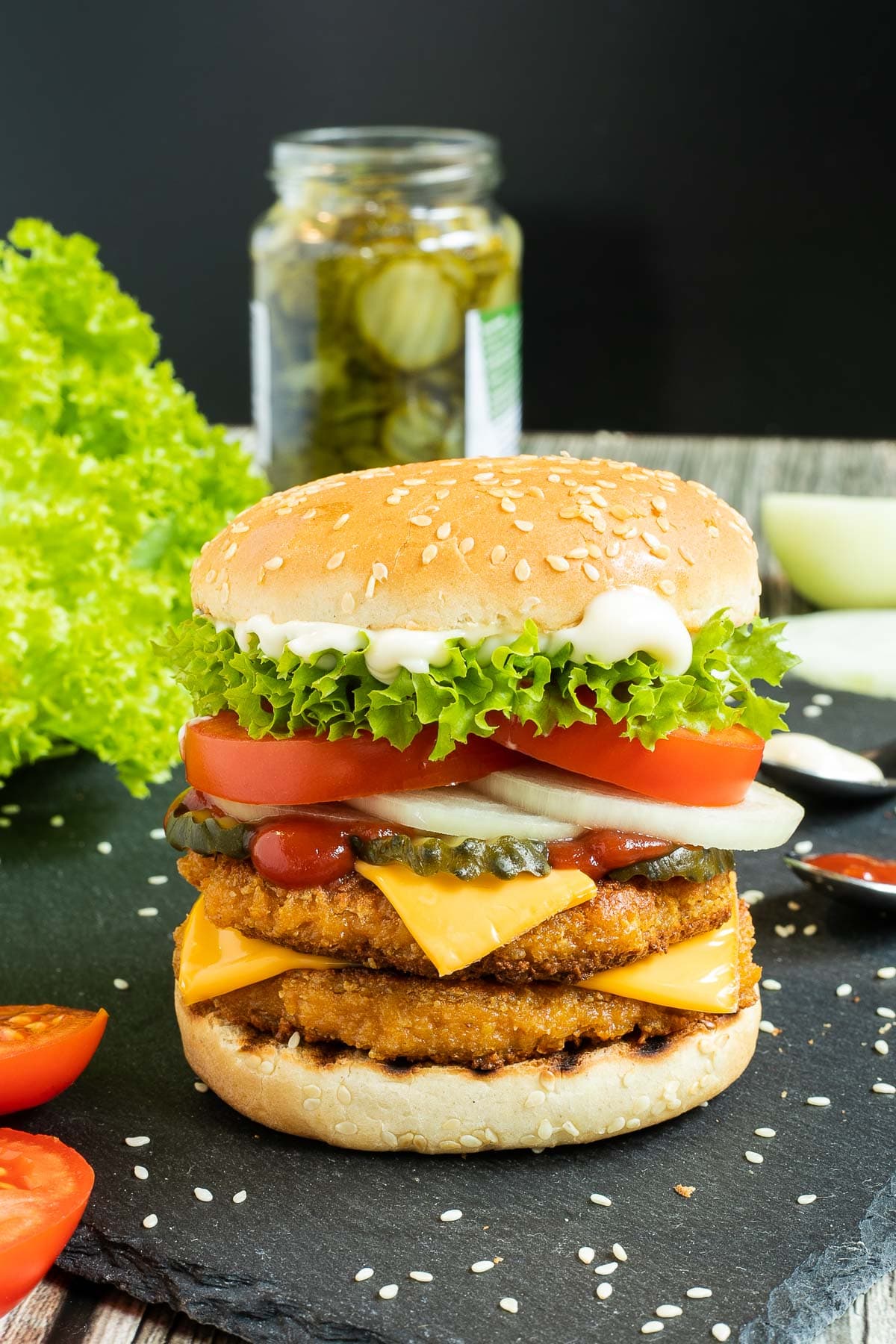 Burger bun with two patties, cheese in between, pickled cucumber slices, onion slices, tomato, and ruffled lettuce. Leftover ingredients are around it like lettuce, jar of pickled cucumber, onion, and tomatoes.