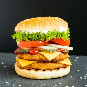 Burger bun with two patties, cheese in between, pickled cucumber slices, onion slices, tomato and ruffled lettuce.