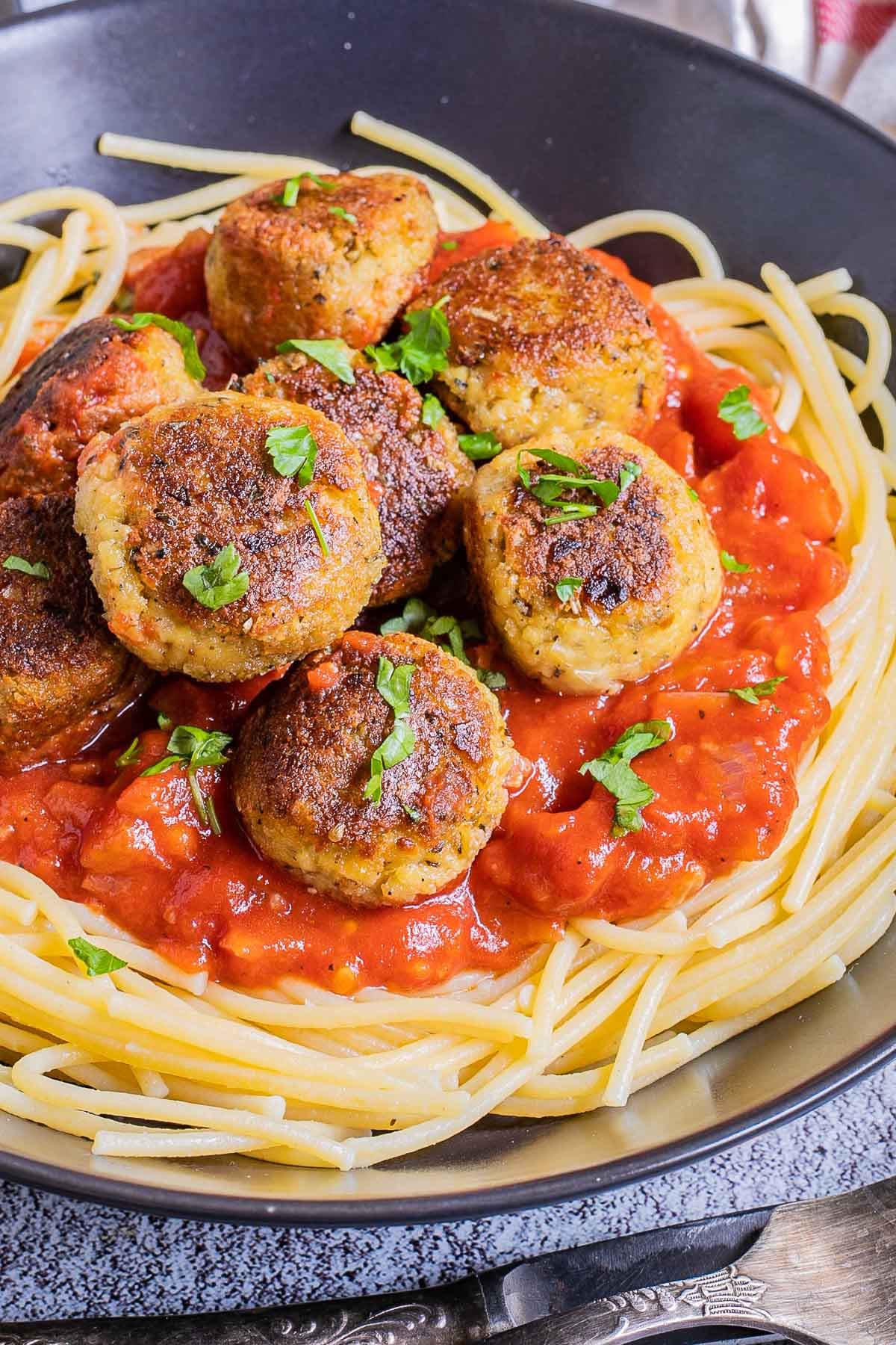 Black plate with spaghetti, marinara sauce and crispy brown tofu meatballs sprinkled with chopped green herbs.
