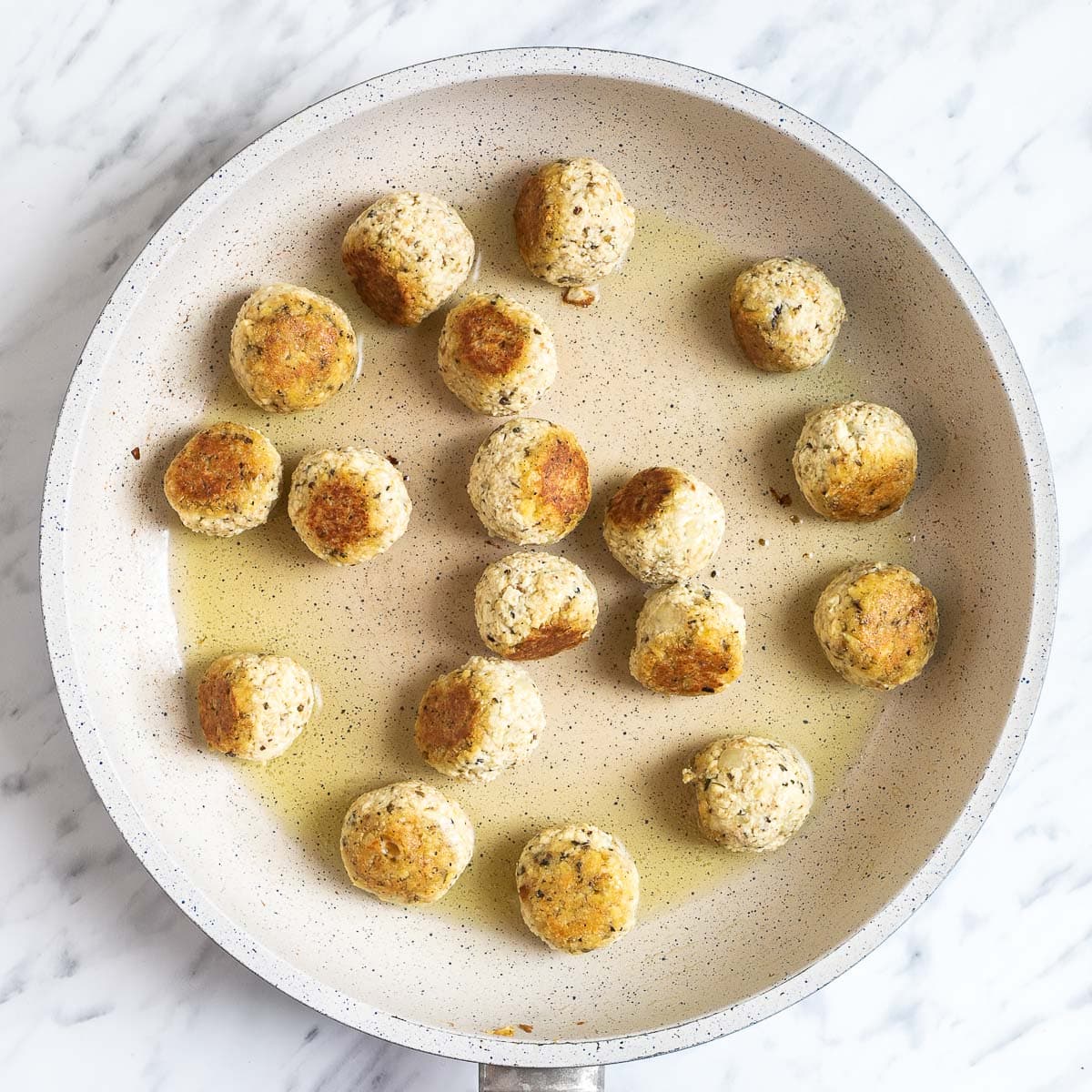 White frying pan with several tofu meatballs getting brownish on one side.