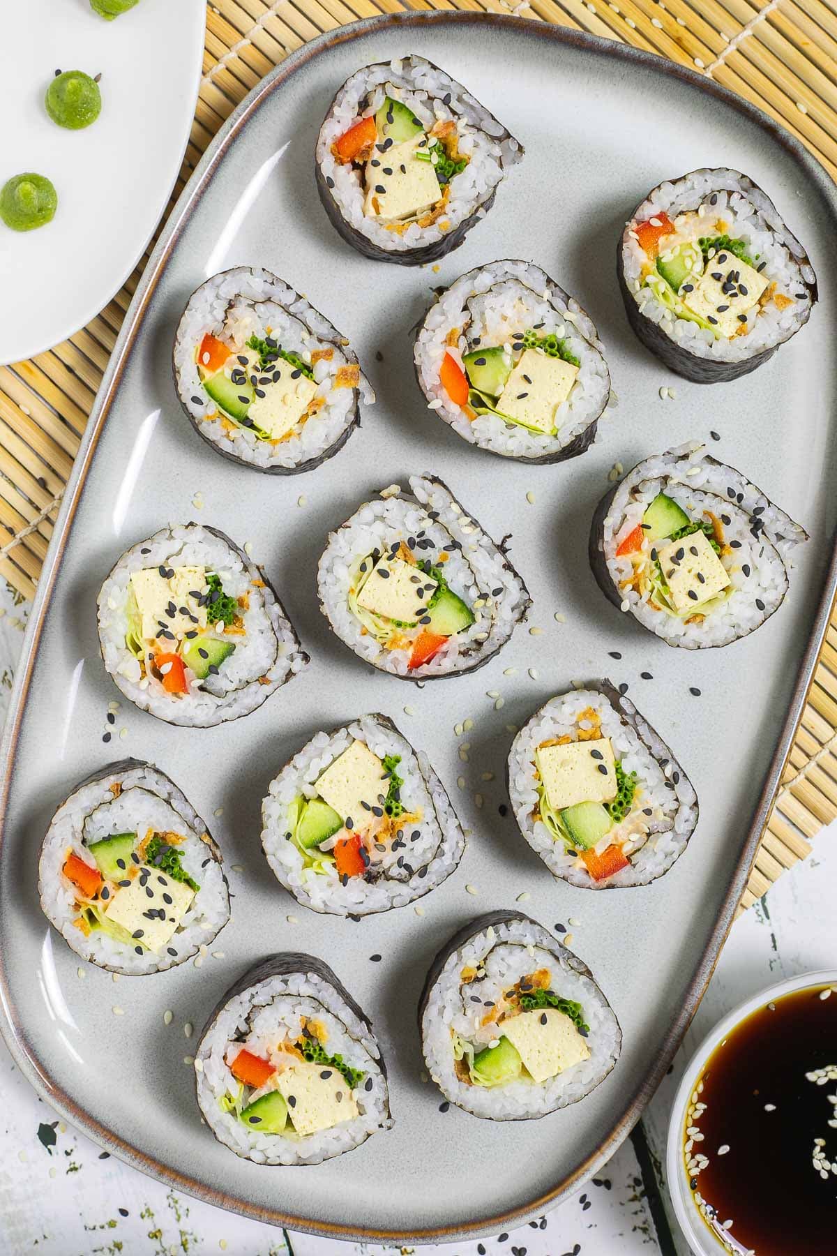 An oval, light blue plate with 9 tofu sushi rolls with colorful filling wrapped in dark seaweed sheets. Served with a small bowl of soy sauce and sprinkled with dark and white sesame seeds.