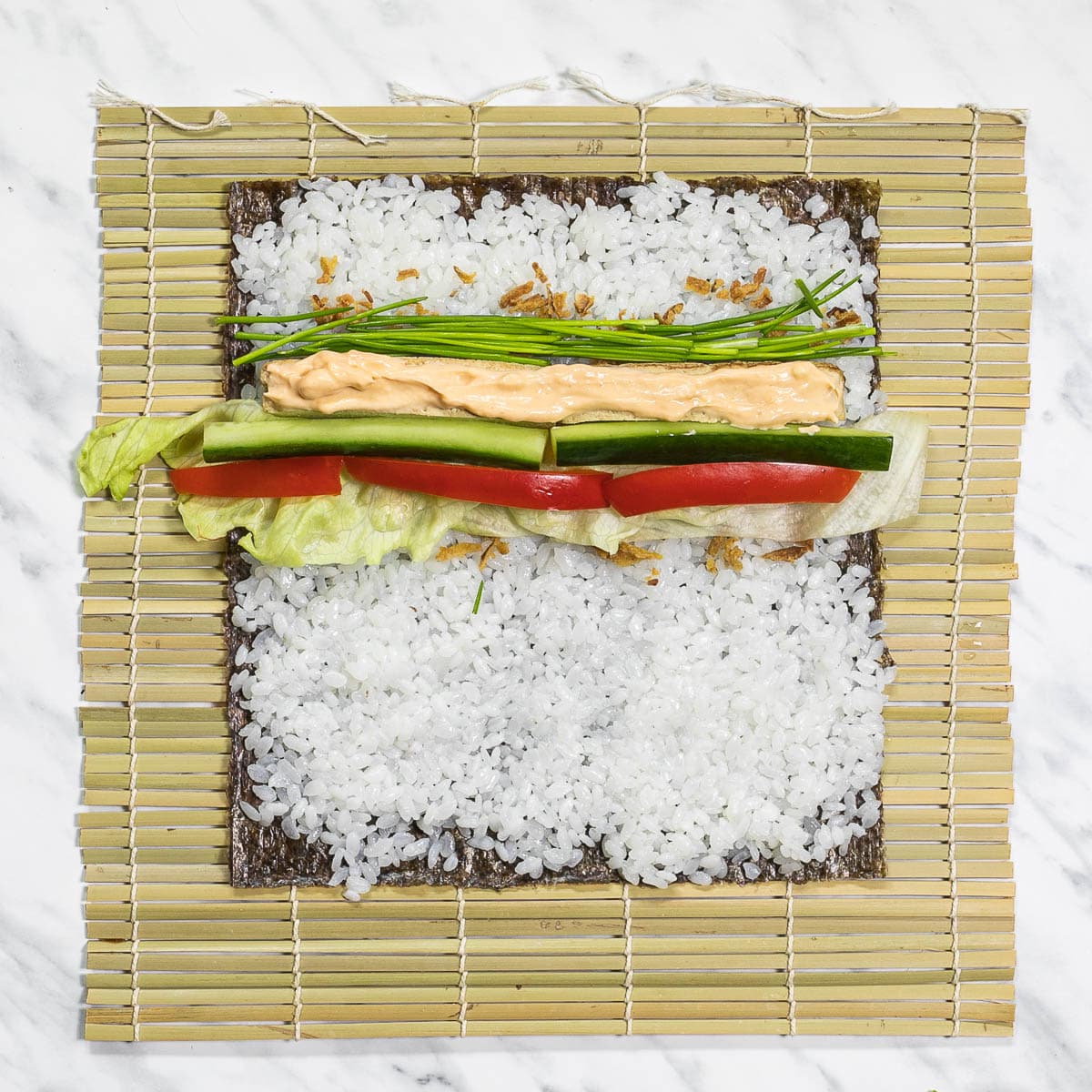 Bamboo mat with seaweed sheets, rice, red pepper, cucumber, chives, and lettuce strips, smoked tofu bar with orange mayo on top. 